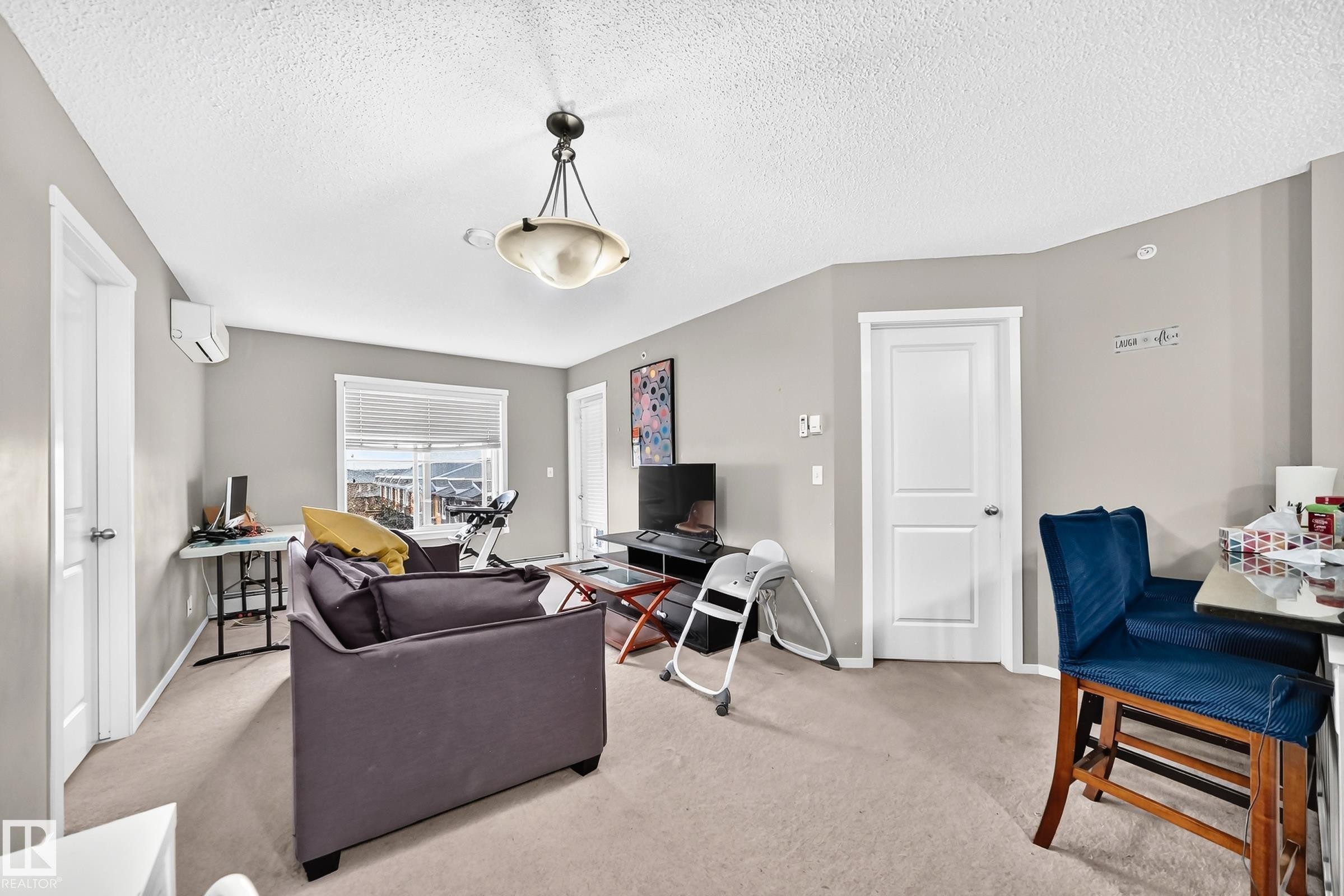 Carpeted common area featuring light gray walls, a window with horizontal blinds, and a built-in air conditioning unit - #417 1510 Watt Dr Sw, Edmonton, AB - Indoor Photo Showing Living Room