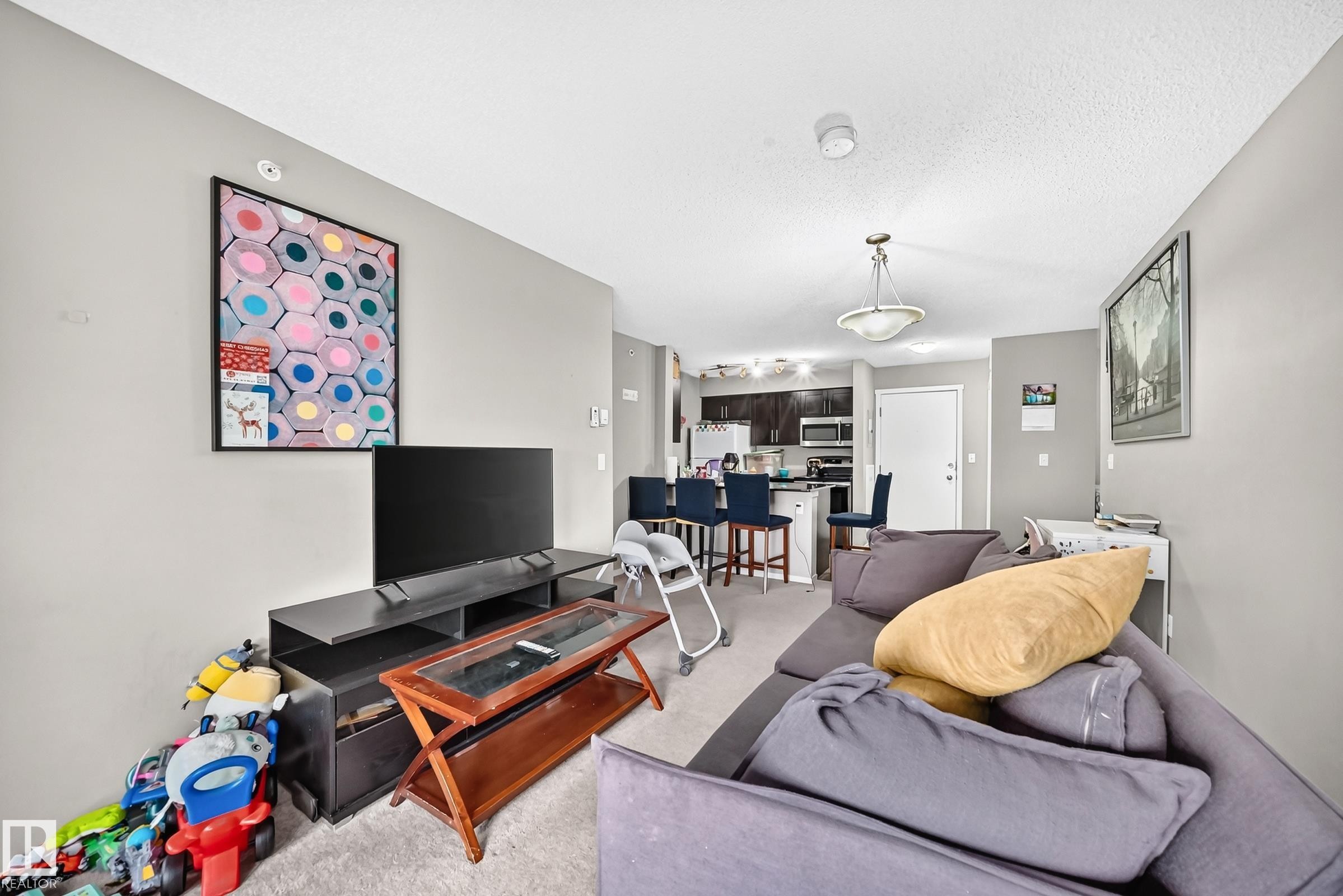 Open-concept living space featuring light gray walls, carpet flooring, and a kitchen area with dark cabinetry, stainless steel appliances, and a breakfast bar - #417 1510 Watt Dr Sw, Edmonton, AB - Indoor