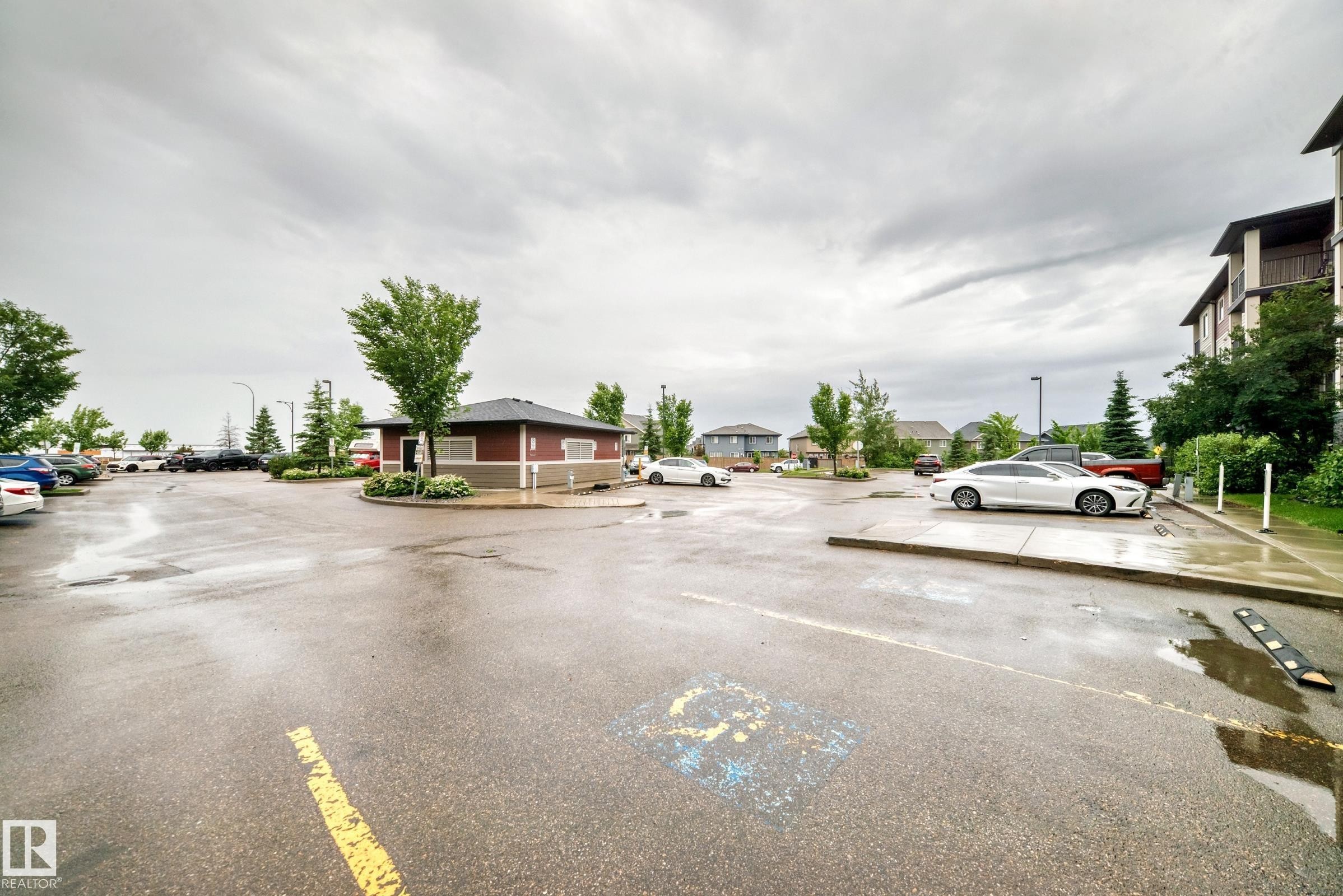 Paved parking area featuring designated accessible parking, a central building with a dark roof and two-tone exterior, and mature landscaping - #417 1510 Watt Dr Sw, Edmonton, AB - Outdoor