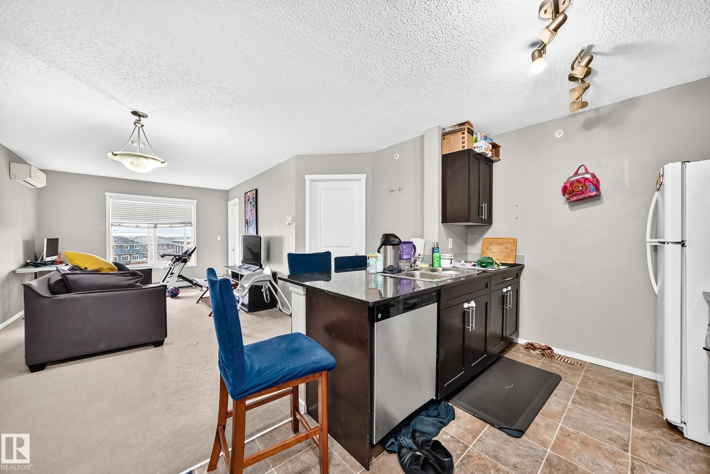 Open-concept living area featuring a kitchen island with dark wood-finish cabinetry, stainless steel dishwasher, and a white refrigerator - #417 1510 Watt Dr Sw, Edmonton, AB - Indoor