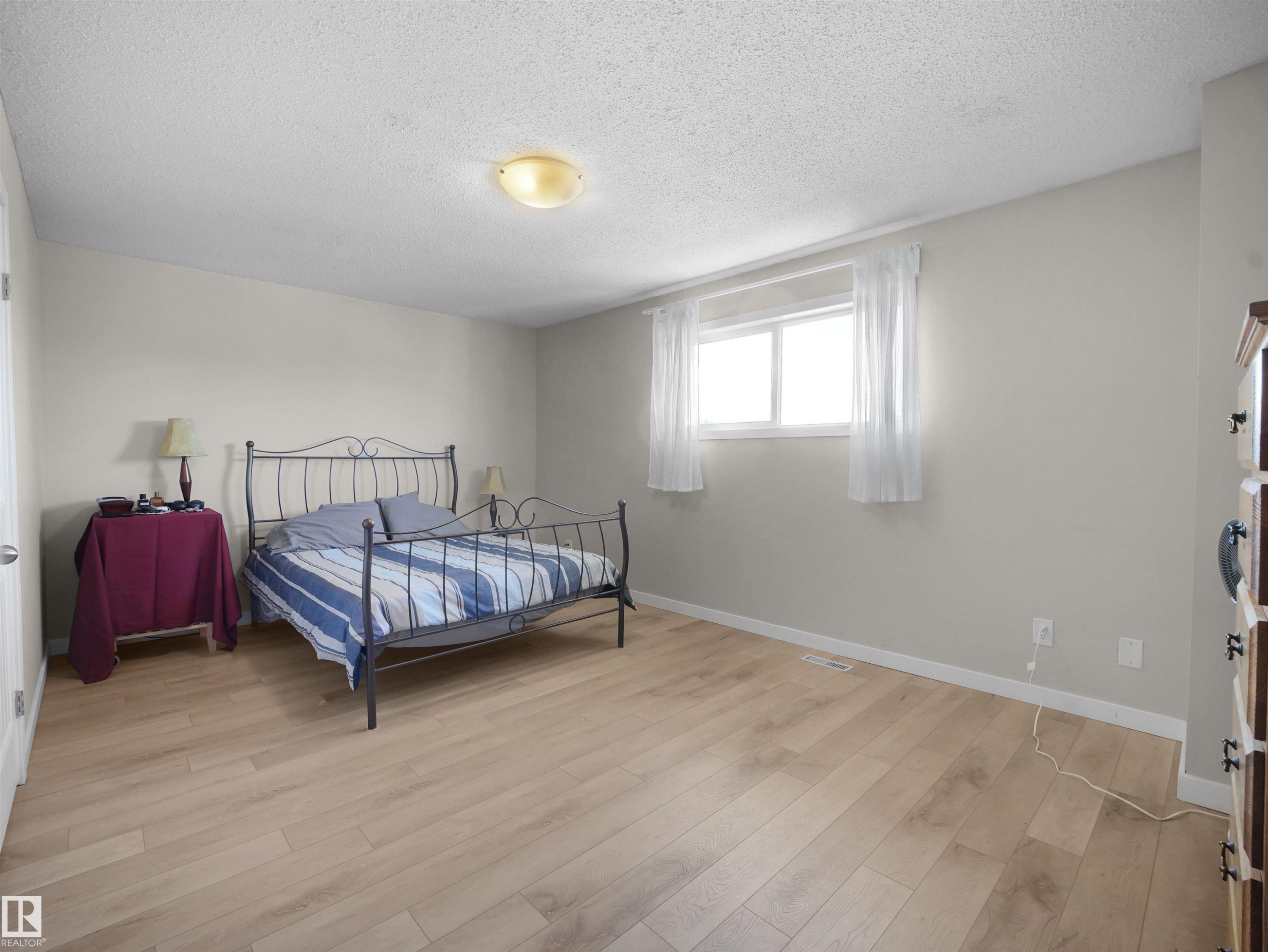 Bright bedroom featuring wood-finish flooring, light neutral wall tones, a ceiling-mounted light fixture, and a white window with sheer curtains - 575 Dunluce Road, Edmonton, AB