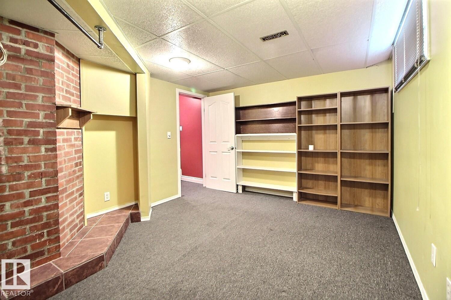 Living space featuring a brick fireplace with a tile hearth, built-in shelving, dropped ceiling, and neutral carpeting - 11312 127 Street Nw, Edmonton, AB