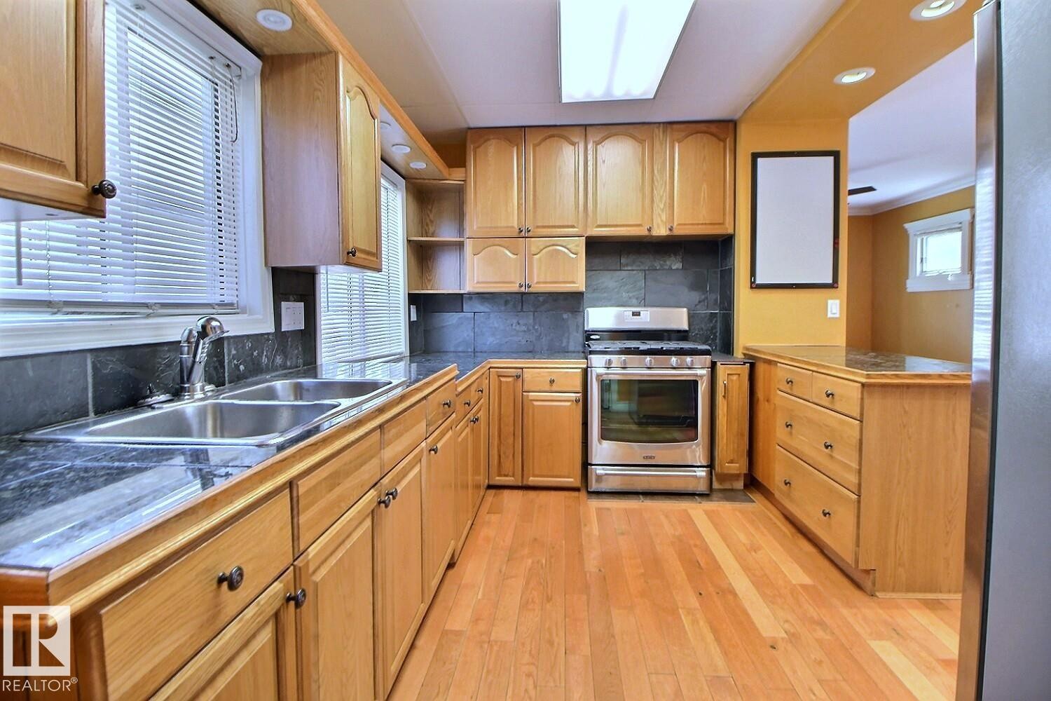Kitchen featuring wood-finish cabinetry, stainless steel appliances, dark stone countertops, a tile backsplash, and light wood-finish flooring - 11312 127 Street Nw, Edmonton, AB