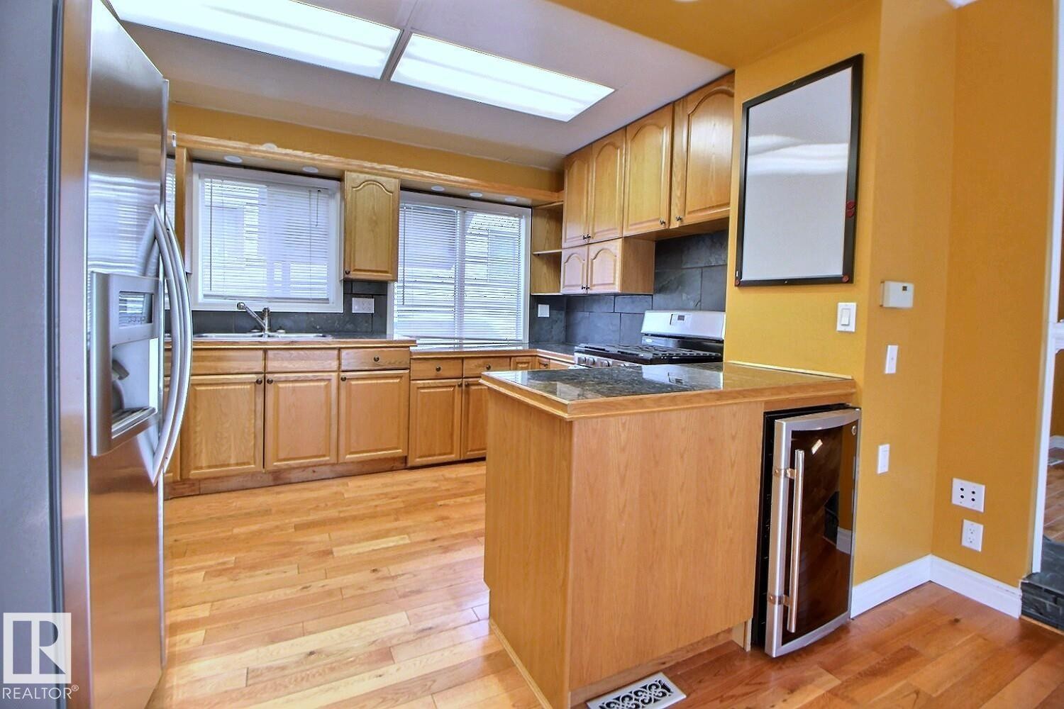 Kitchen featuring wood-finish flooring, natural wood cabinetry, a stainless steel refrigerator, a built-in beverage cooler, and a dark stone countertop - 11312 127 Street Nw, Edmonton, AB