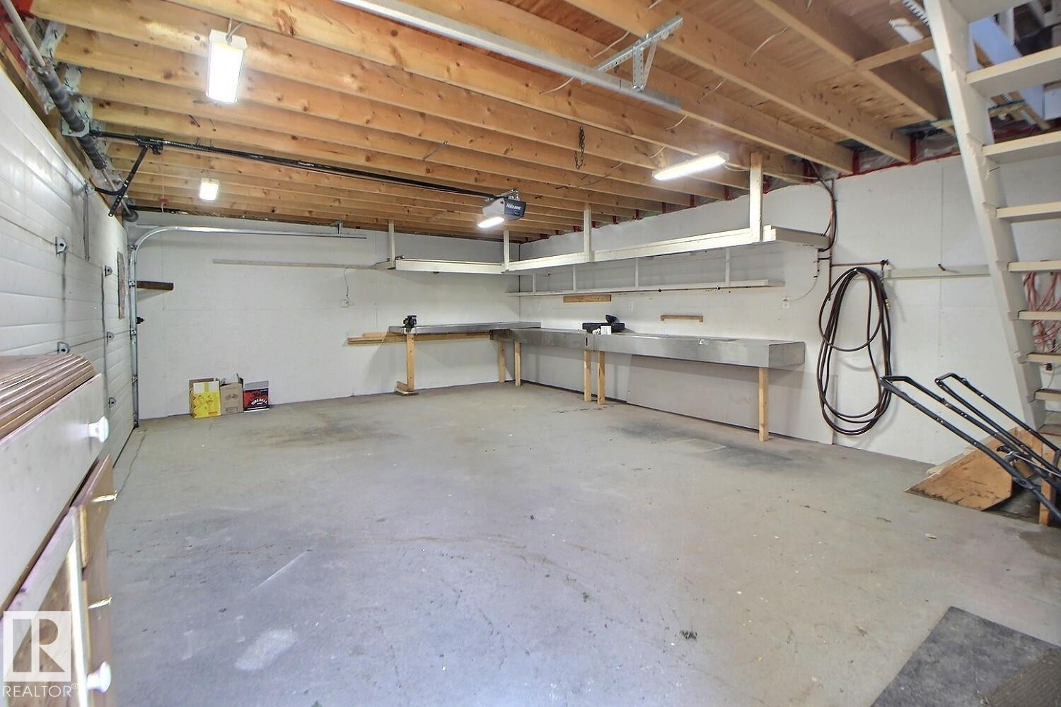 Spacious garage featuring exposed wood ceiling joists, white insulated garage door, concrete flooring, built-in shelving, and extensive workbench space - 11312 127 Street Nw, Edmonton, AB