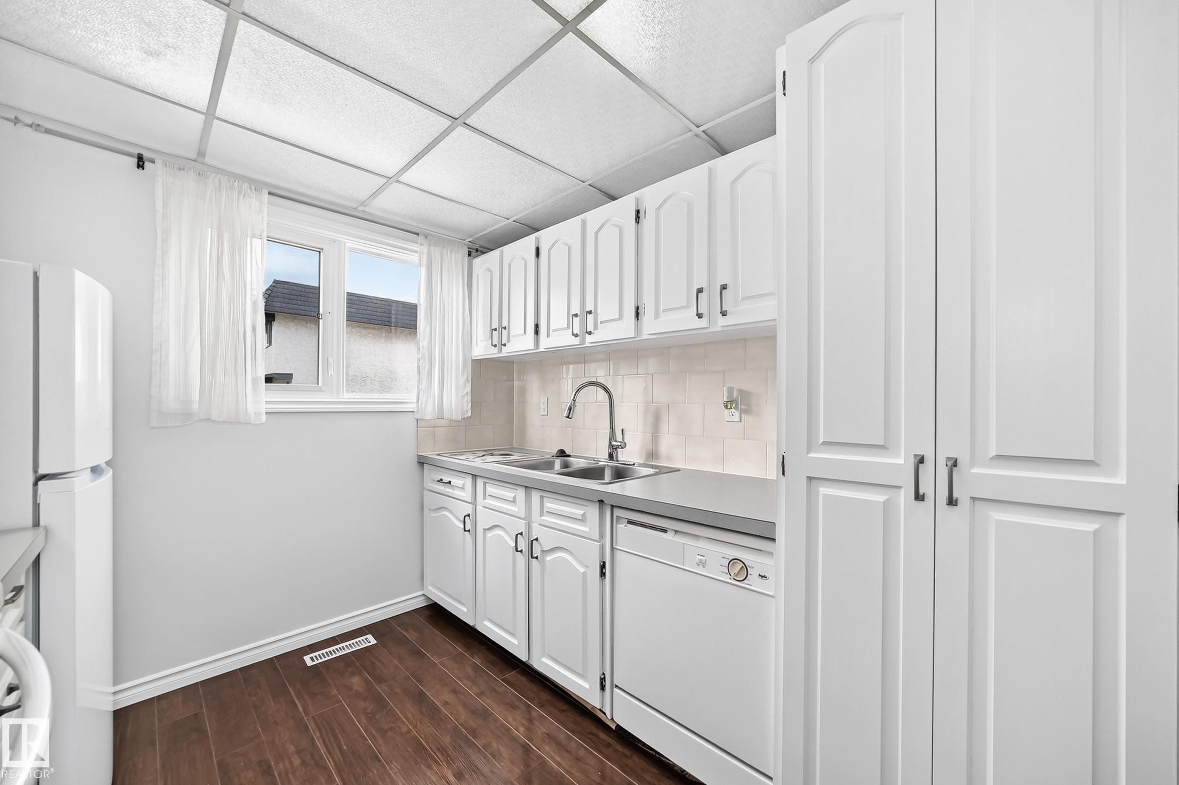 Kitchen featuring white cabinetry, dark wood-finish flooring, a double basin sink with a gooseneck faucet, a light-toned countertop, and a tiled backsplash - 11447 22 Avenue Nw, Edmonton, AB - Indoor Photo Showing Kitchen With Double Sink