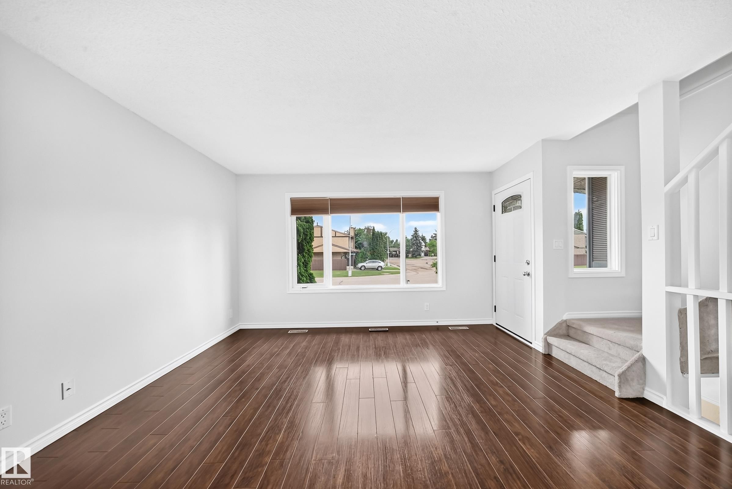 Spacious living area featuring rich wood-finish flooring, a large picture window with wood blinds, and crisp white baseboards - 11447 22 Avenue Nw, Edmonton, AB - Indoor