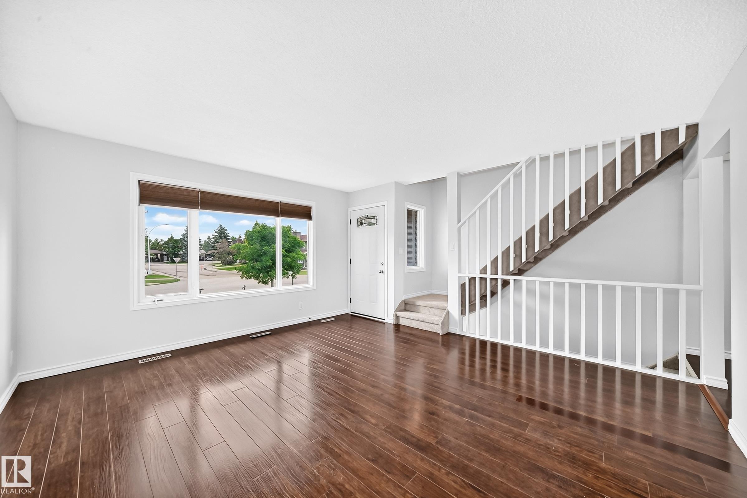 Spacious living area featuring wood-finish flooring, a large picture window with blinds, and white painted walls - 11447 22 Avenue Nw, Edmonton, AB - Indoor