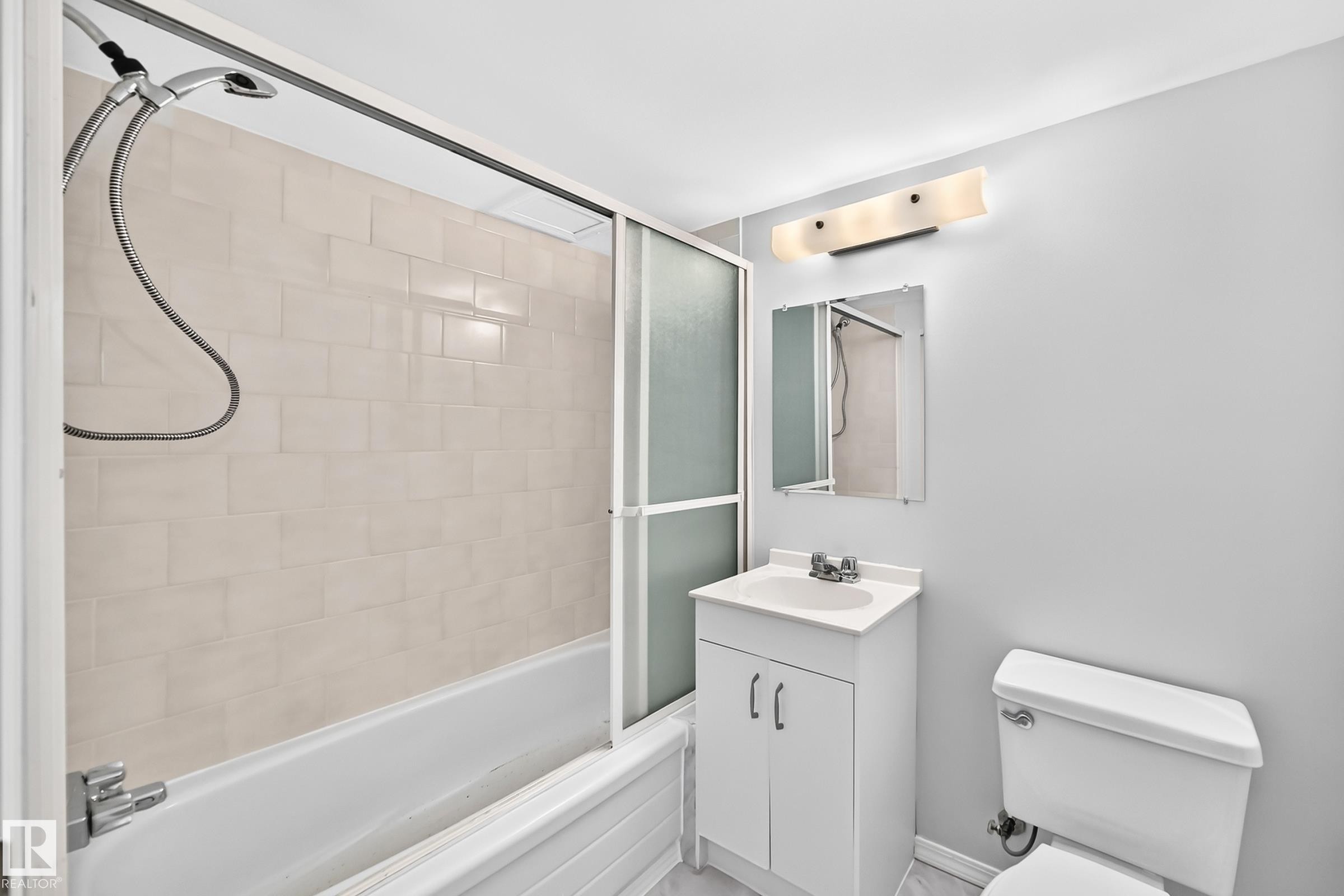 Bathroom featuring a white bathtub with tile surround, sliding frosted glass shower doors, a vanity with integrated sink, a wall-mounted mirror, and a modern light fixture - 11447 22 Avenue Nw, Edmonton, AB - Indoor Photo Showing Bathroom