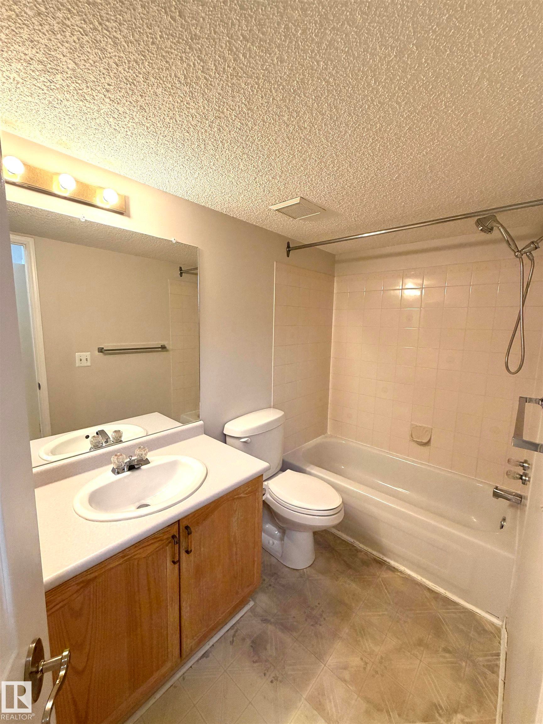 Bathroom featuring a vanity with wood-finish cabinetry and an integrated sink, a full-width mirror, a toilet, and a bathtub with tile surround - 207 18012 95 Avenue, Edmonton, AB