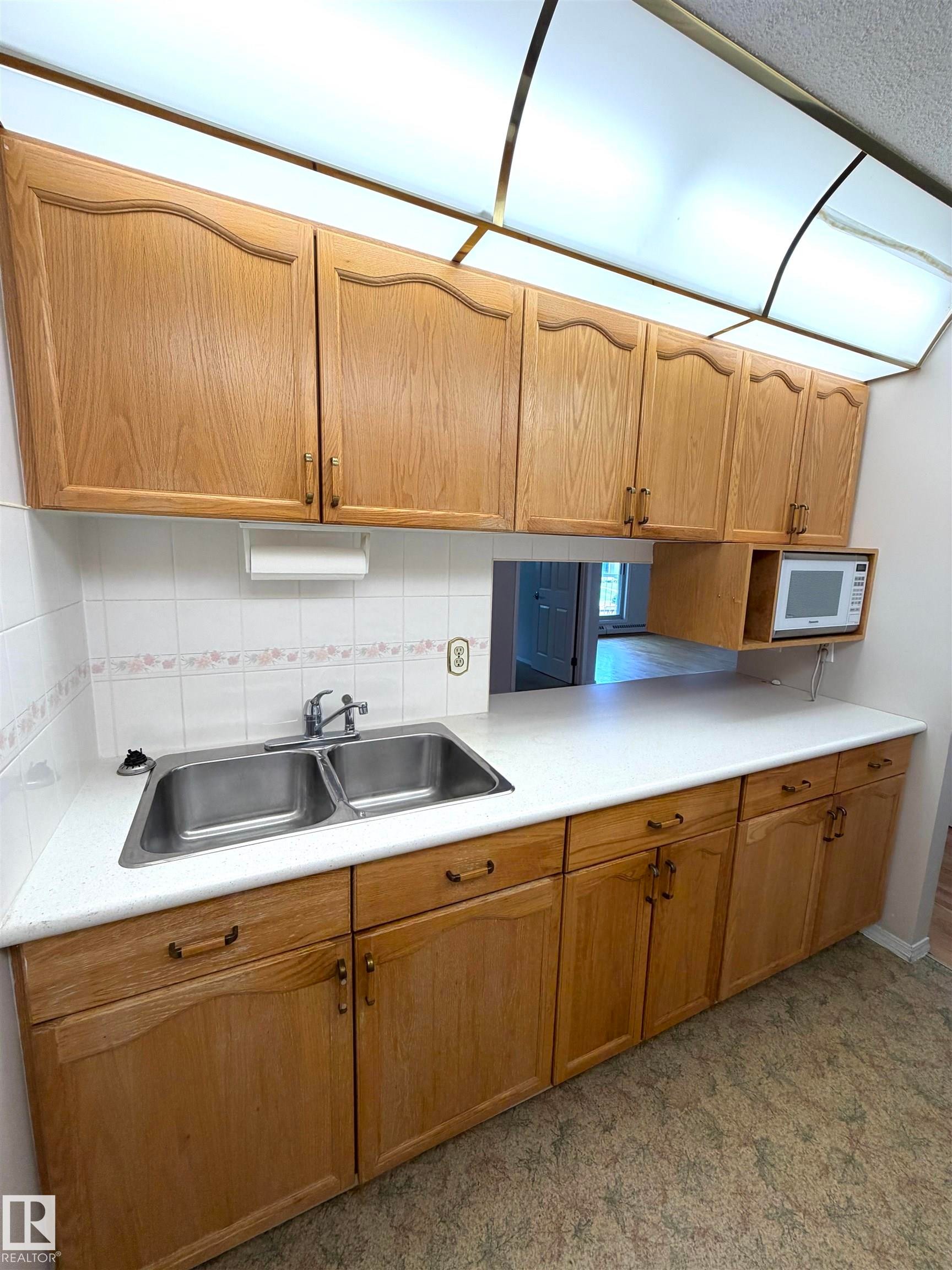 Kitchen featuring wood-finish cabinetry, a double basin stainless steel sink, a microwave alcove, white countertops, and a white tile backsplash - 207 18012 95 Avenue, Edmonton, AB