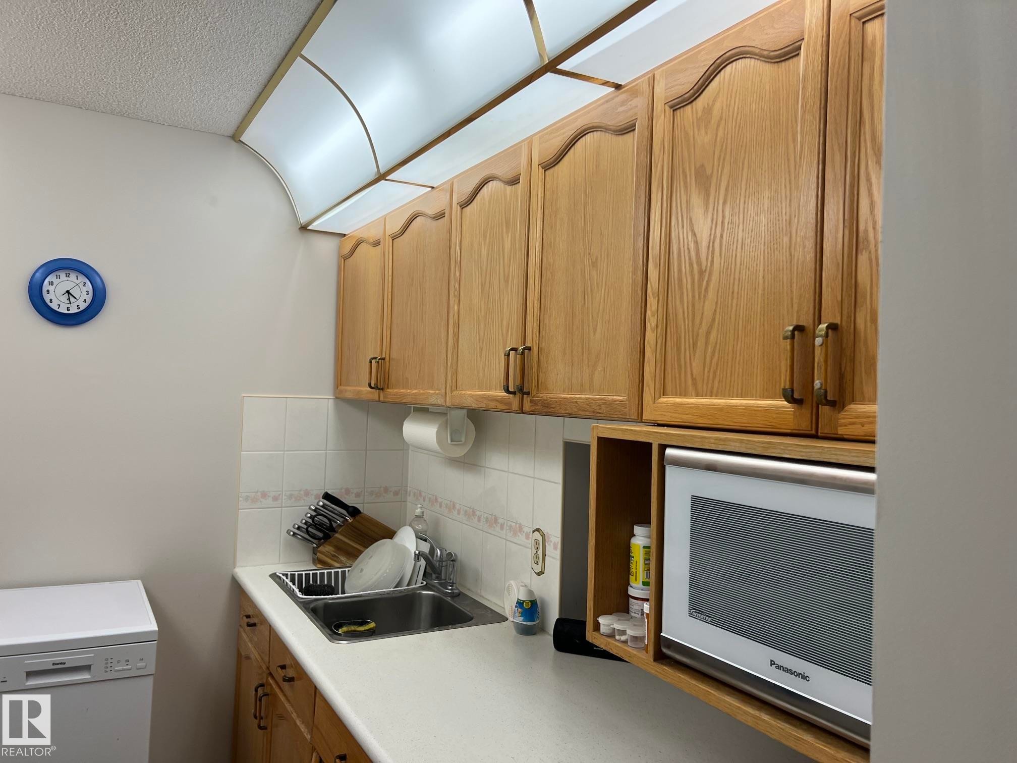 Kitchenette featuring wood-finish cabinetry, built-in microwave, white tile backsplash, a single basin sink, and an integrated overhead light fixture - 207 18012 95 Avenue, Edmonton, AB