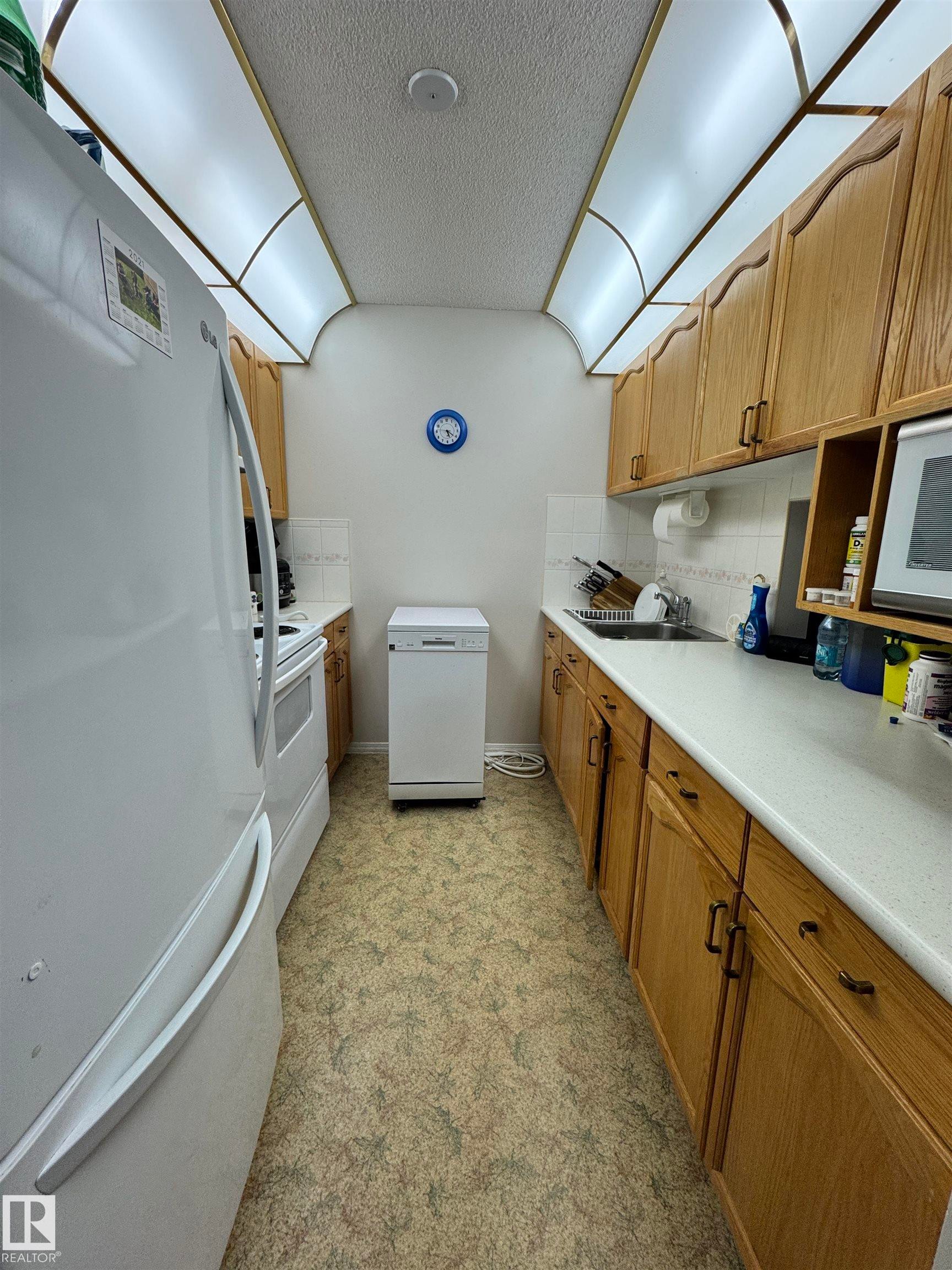 Galley kitchen featuring wood cabinetry, white laminate countertops, and a white tile backsplash - 207 18012 95 Avenue, Edmonton, AB