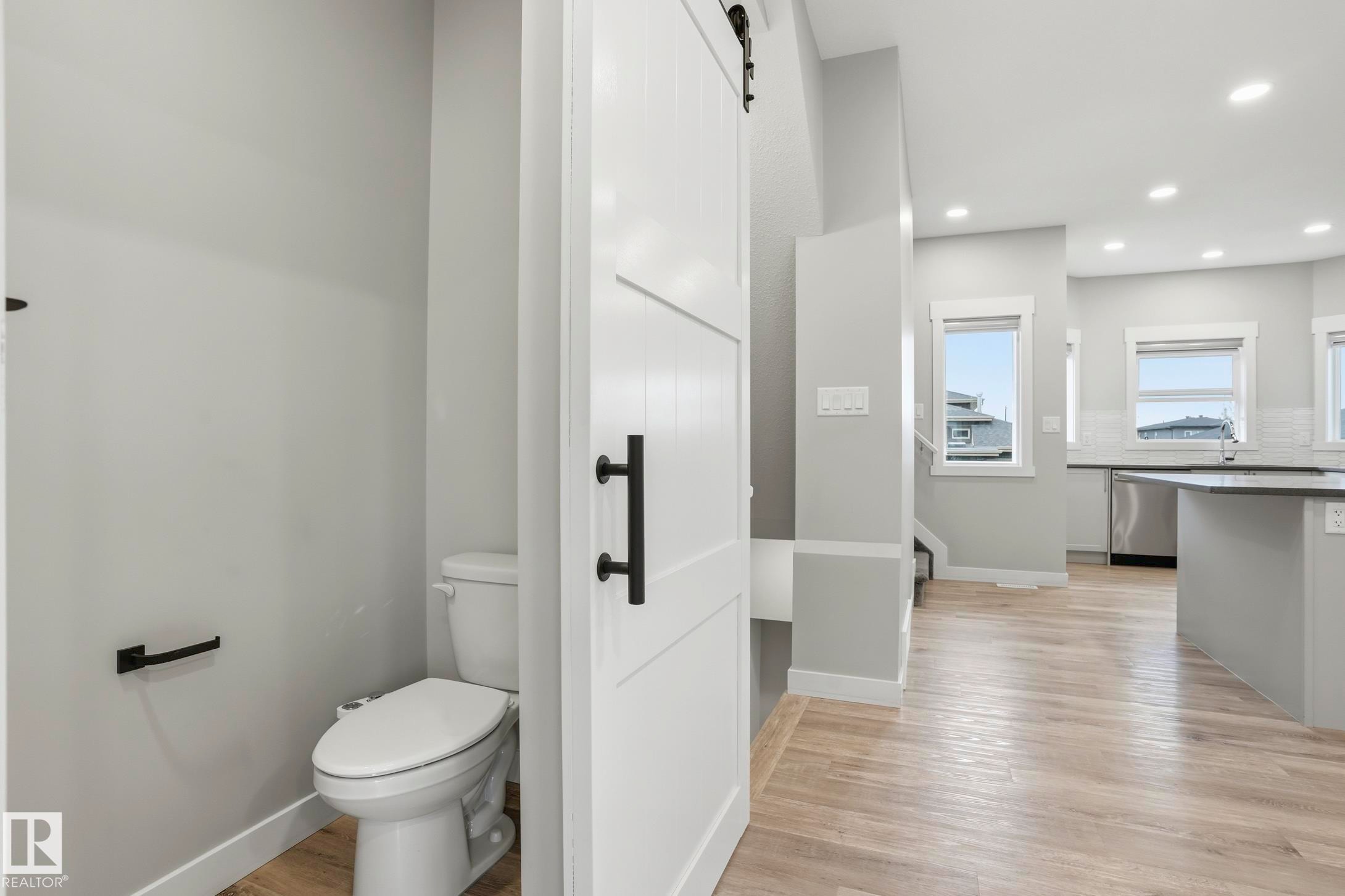 White barn door entryway with black hardware, wood-finish flooring, and a powder room featuring a white toilet - 36 150 Everitt Dr, St. Albert, AB - Indoor Photo Showing Bathroom
