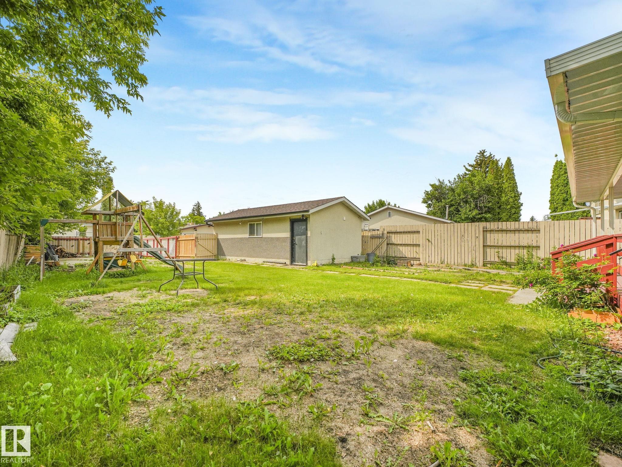 Expansive backyard with a mix of grass and soil, featuring a detached structure with a stucco and siding exterior, a wood fence, and mature trees providing natural screening - 9315 151 Avenue, Edmonton, AB