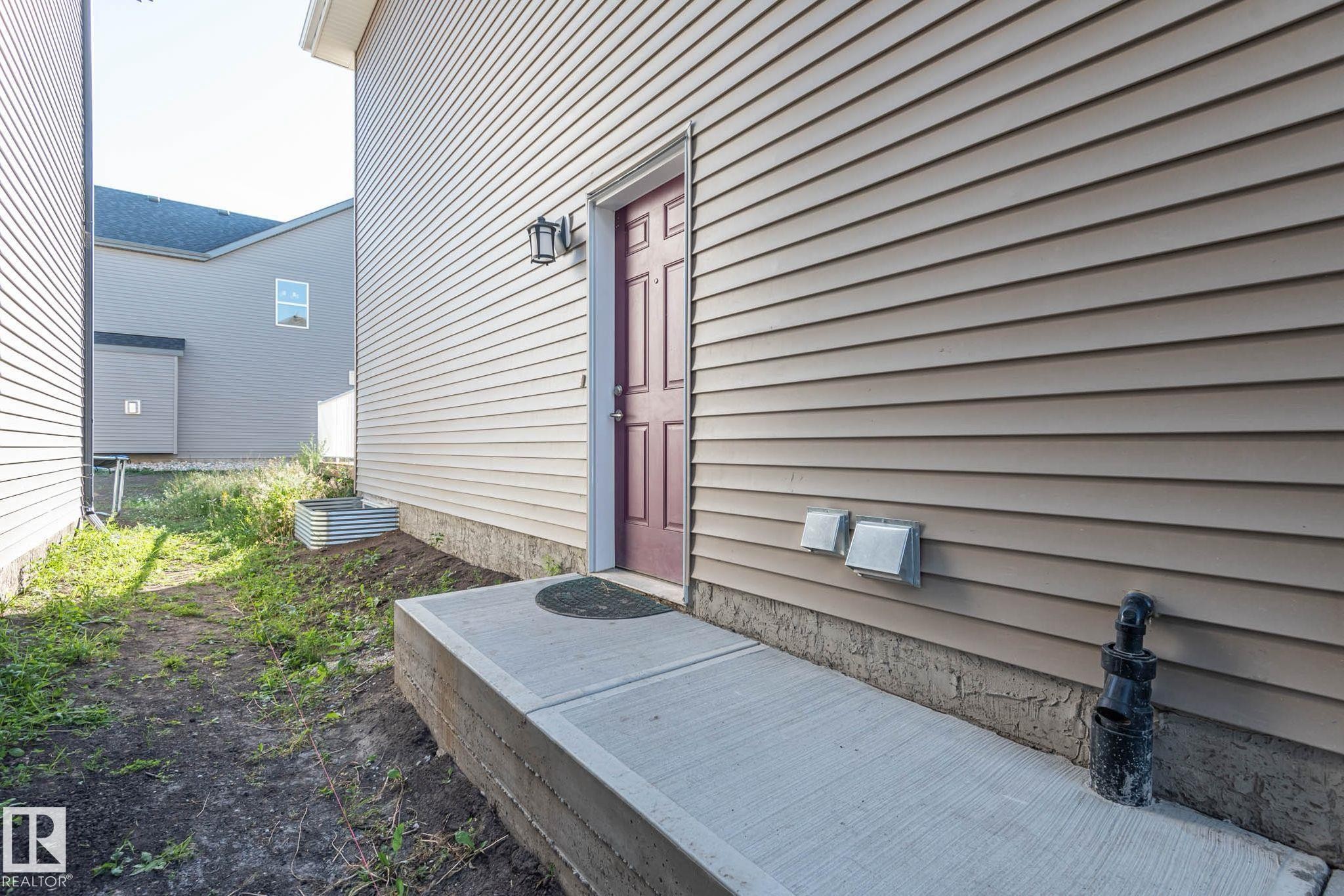Exterior side entrance with a concrete walkway, light-toned horizontal siding, and a red paneled door - 2635 Donaghey Crescent, Edmonton, AB