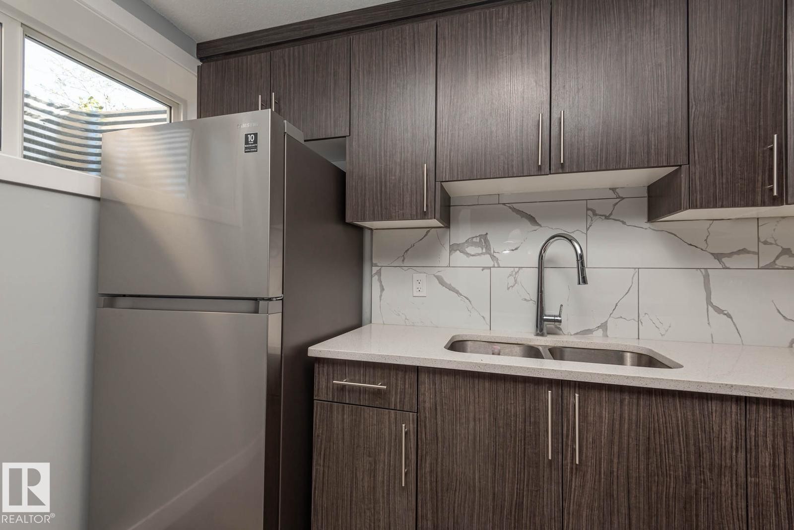 Contemporary kitchen featuring dark wood-finish cabinetry, a stainless steel refrigerator, white quartz-finish countertops, a dual basin stainless steel sink with a gooseneck faucet, and a large format tile backsplash with a grey vein pattern - 2635 Donaghey Crescent, Edmonton, AB