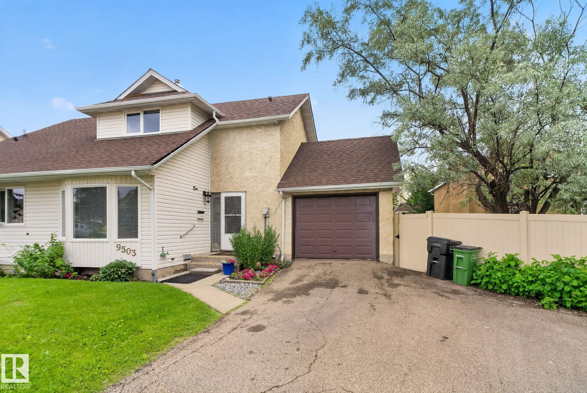 Exterior featuring light-colored siding and stucco finishes, a prominent bay window, an integrated single-car garage with a brown door, a front entry with a small porch, and a dormer window - 9503 180A Street Nw, Edmonton, AB - Outdoor