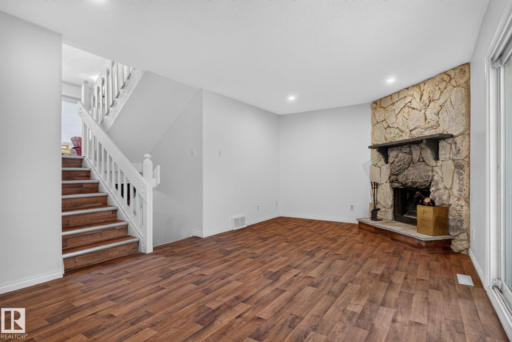 Living area featuring a stone fireplace with a dark mantel, wood-finish flooring, recessed lighting, white walls, and a staircase with white balusters and wood-finish treads - 9503 180A Street Nw, Edmonton, AB - Indoor Photo Showing Other Room With Fireplace