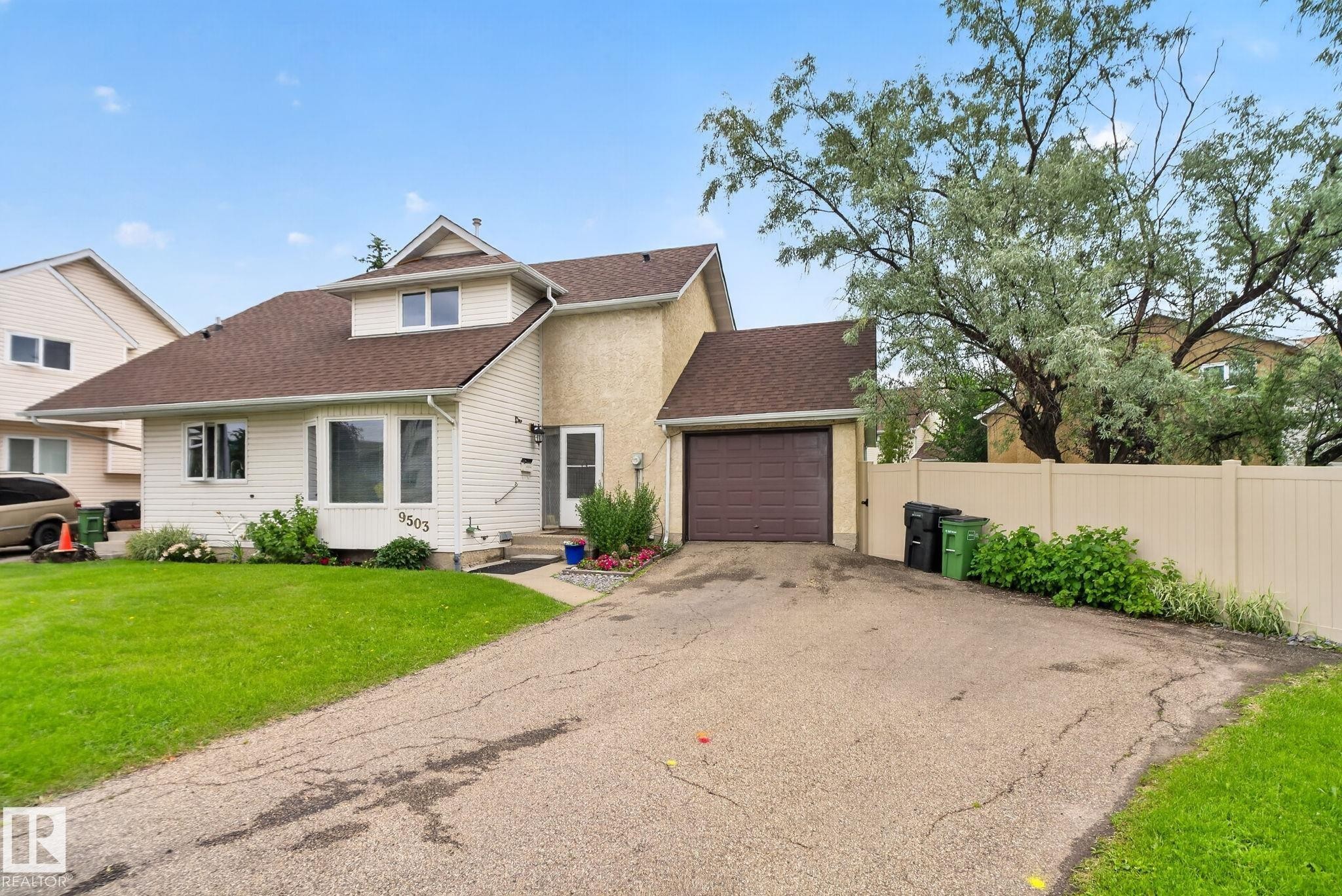 Two-story residence featuring a combination of cream stucco and white siding, a brown shingle roof, an attached garage with a brown door, a front lawn, and a light-colored privacy fence - 9503 180A Street Nw, Edmonton, AB - Outdoor