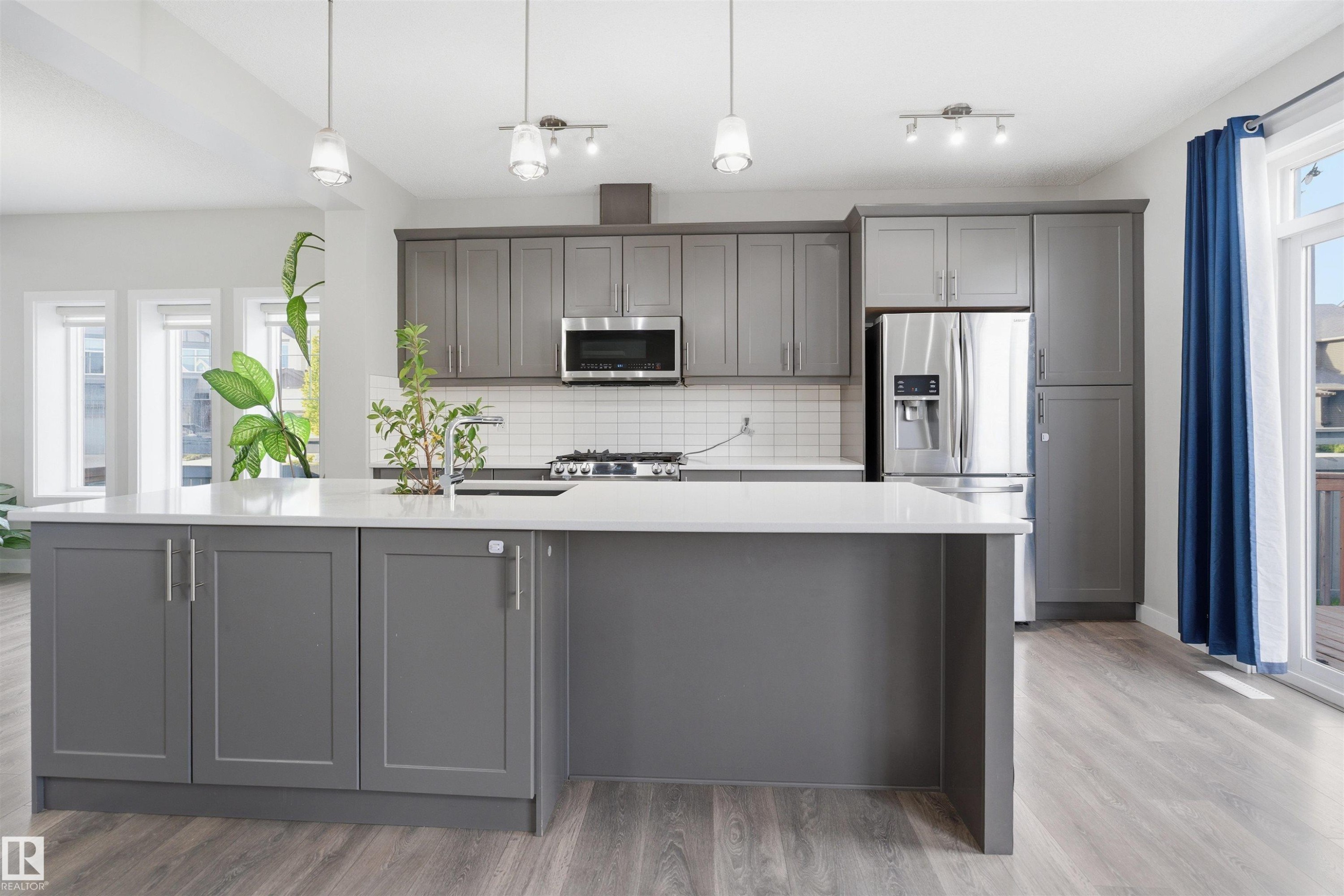 Contemporary kitchen featuring an expansive central island with a solid surface countertop, grey shaker-style cabinetry, stainless steel appliances, and wood-finish flooring - 1234 Daniels Crescent, Edmonton, AB - Indoor Photo Showing Kitchen With Stainless Steel Kitchen With Upgraded Kitchen