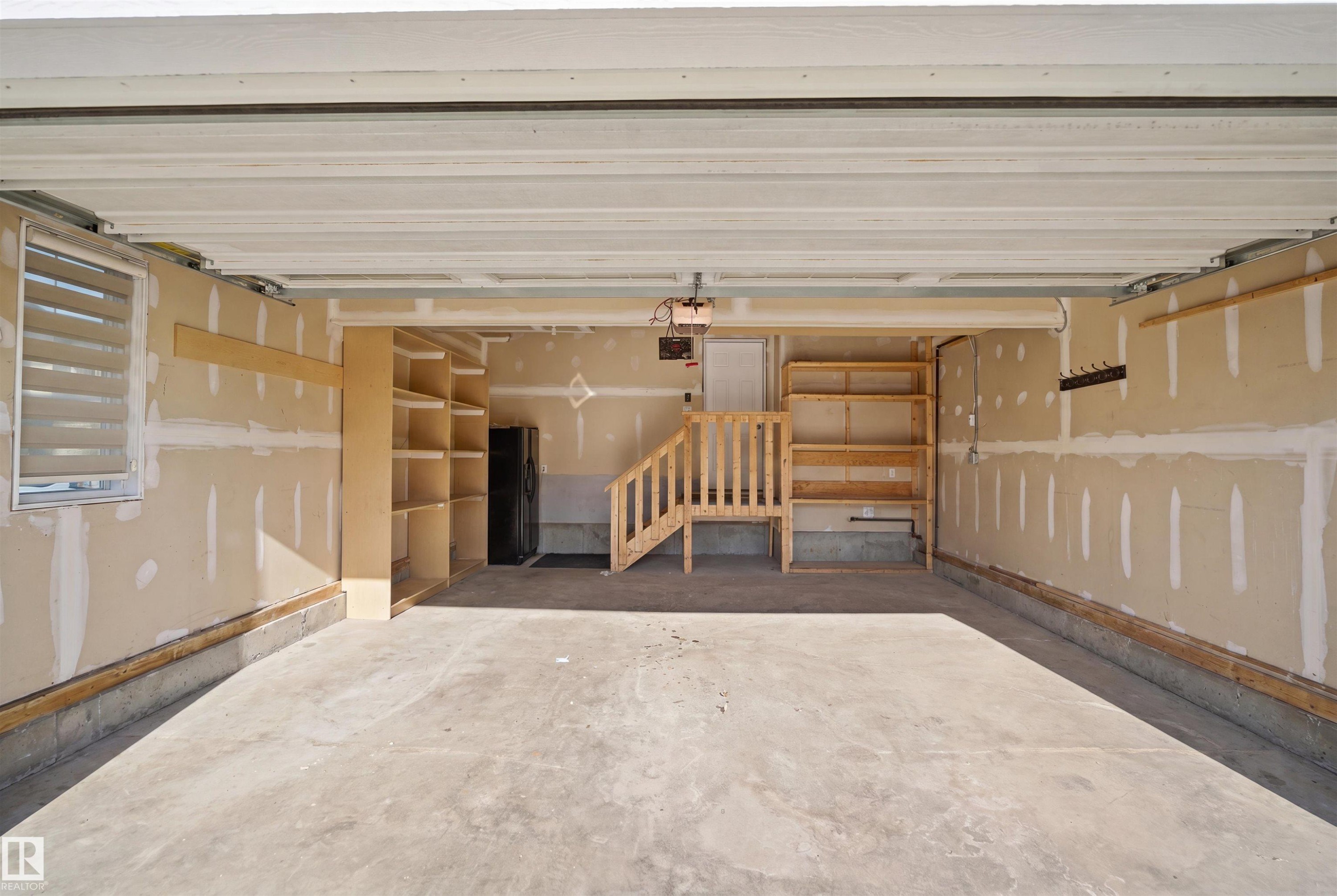 Spacious garage featuring a concrete floor, built-in shelving, a window with horizontal blinds, and a utility staircase - 1234 Daniels Crescent, Edmonton, AB - Photo Showing Garage