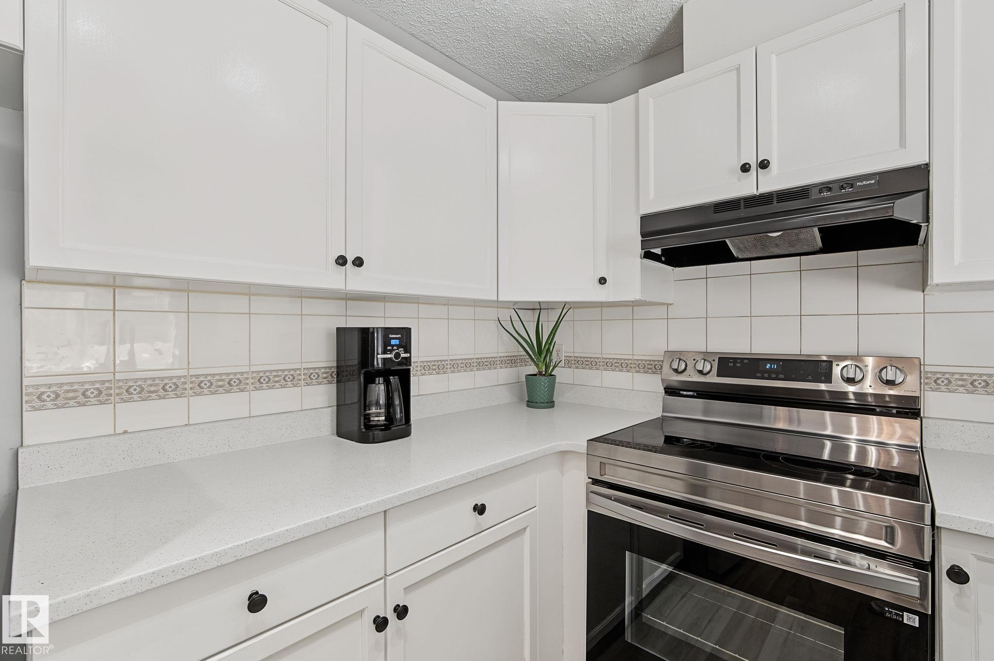 Kitchen featuring white cabinetry, solid surface countertops, white tile backsplash with decorative accent, and a stainless steel range with a built-in exhaust hood - 5 14603 Miller Boulevard, Edmonton, AB
