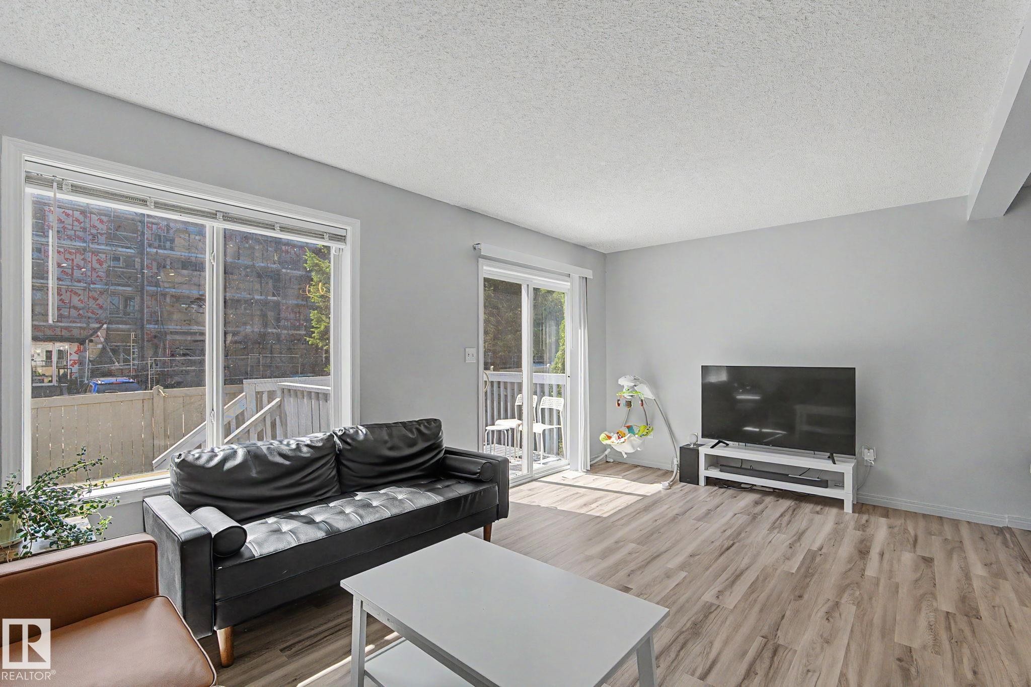 Living area featuring light gray walls, wood-finish flooring, a large window, and a sliding glass door opening to an exterior deck - 5 14603 Miller Boulevard, Edmonton, AB