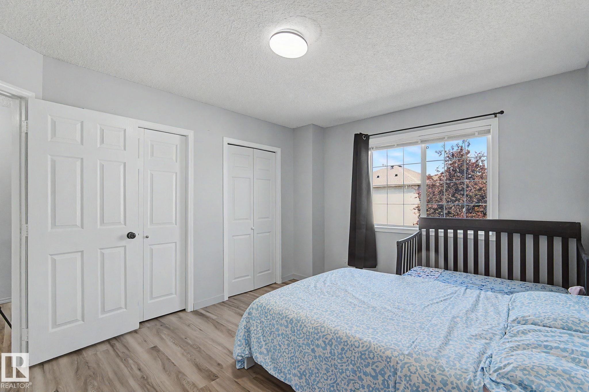 Bedroom featuring light wood-finish flooring, light gray wall paint, a window with exterior residential visibility, and two white bi-fold closet doors - 5 14603 Miller Boulevard, Edmonton, AB