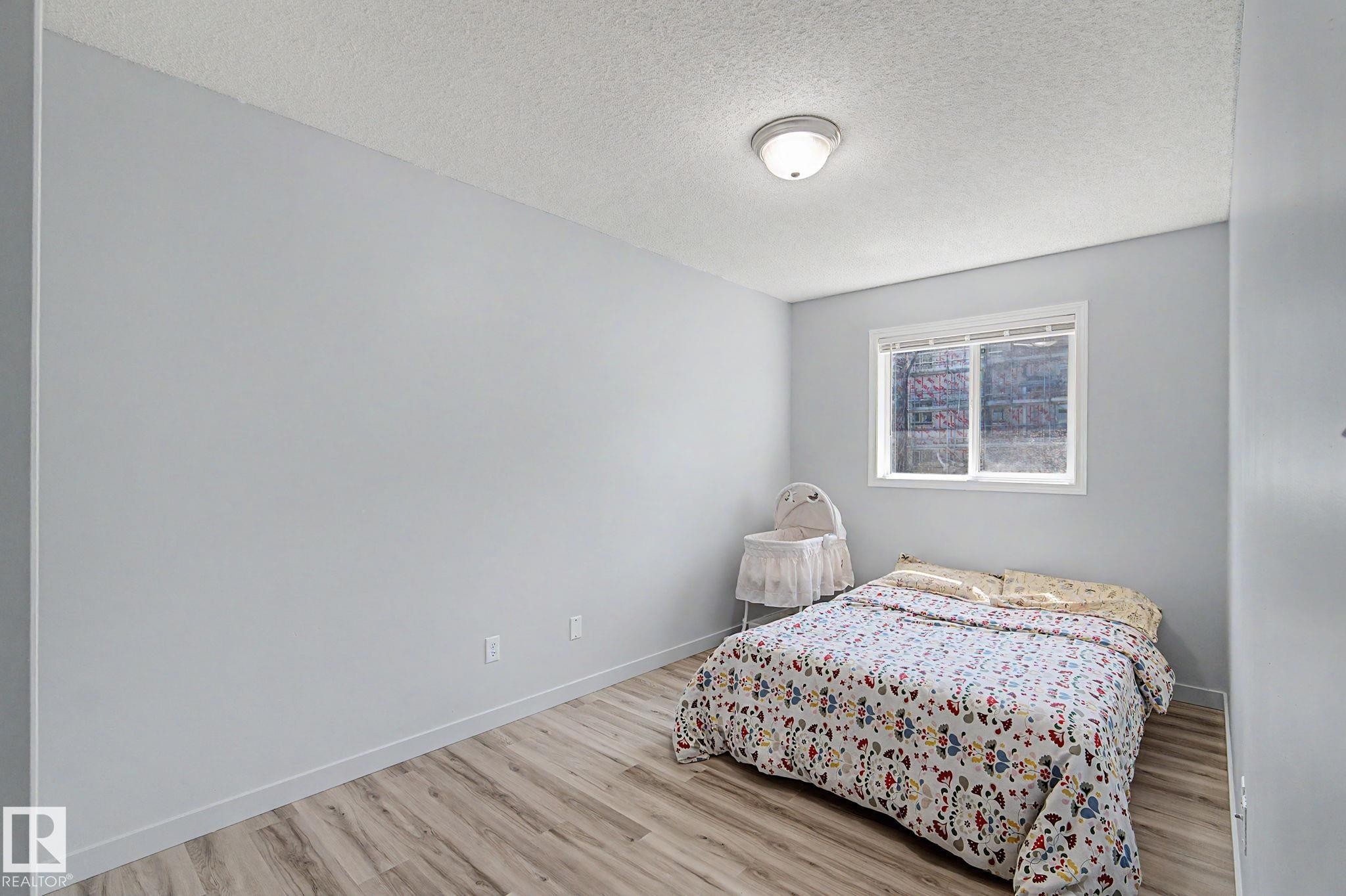 Bedroom featuring light gray walls, wood-finish flooring, and a white-framed window - 5 14603 Miller Boulevard, Edmonton, AB