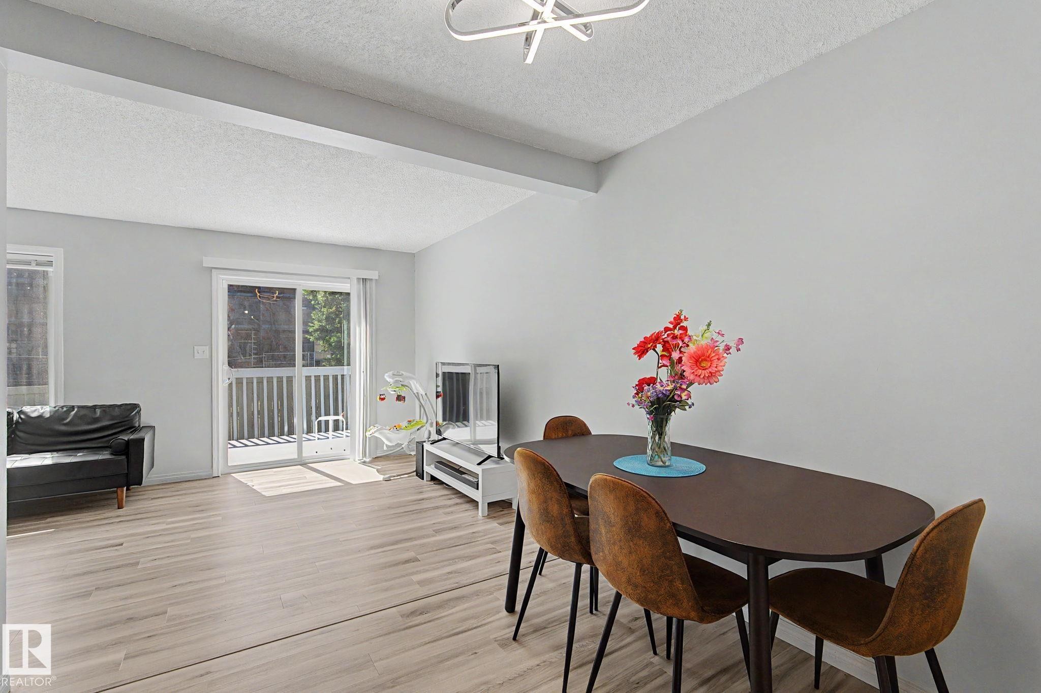 Open-concept living area featuring light wood-finish flooring, a modern flush-mount light fixture, and a sliding glass door leading to an outdoor balcony - 5 14603 Miller Boulevard, Edmonton, AB