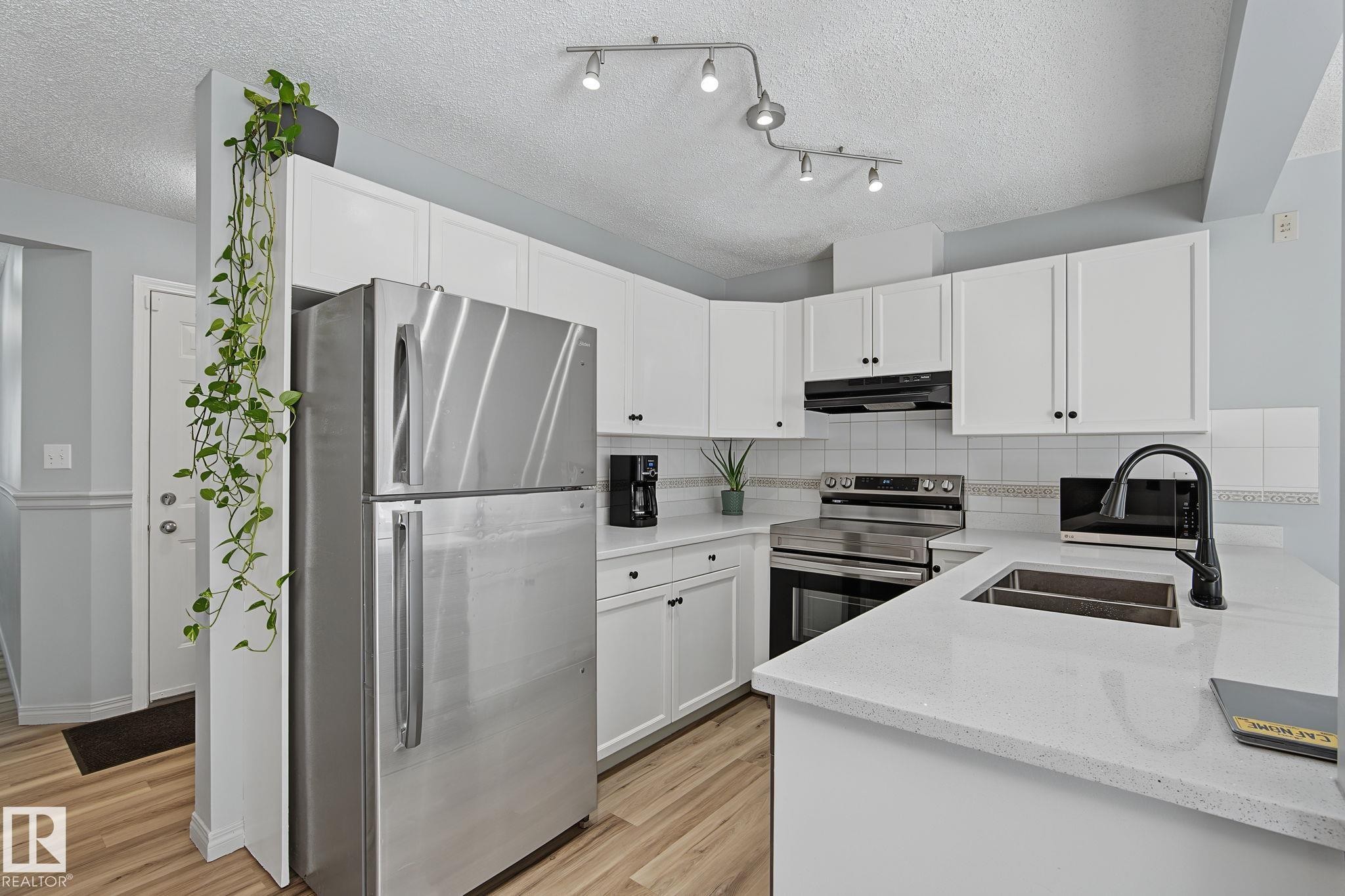 Contemporary kitchen featuring white cabinetry, light-toned countertops, stainless steel appliances, a black faucet, and wood-finish flooring - 5 14603 Miller Boulevard, Edmonton, AB