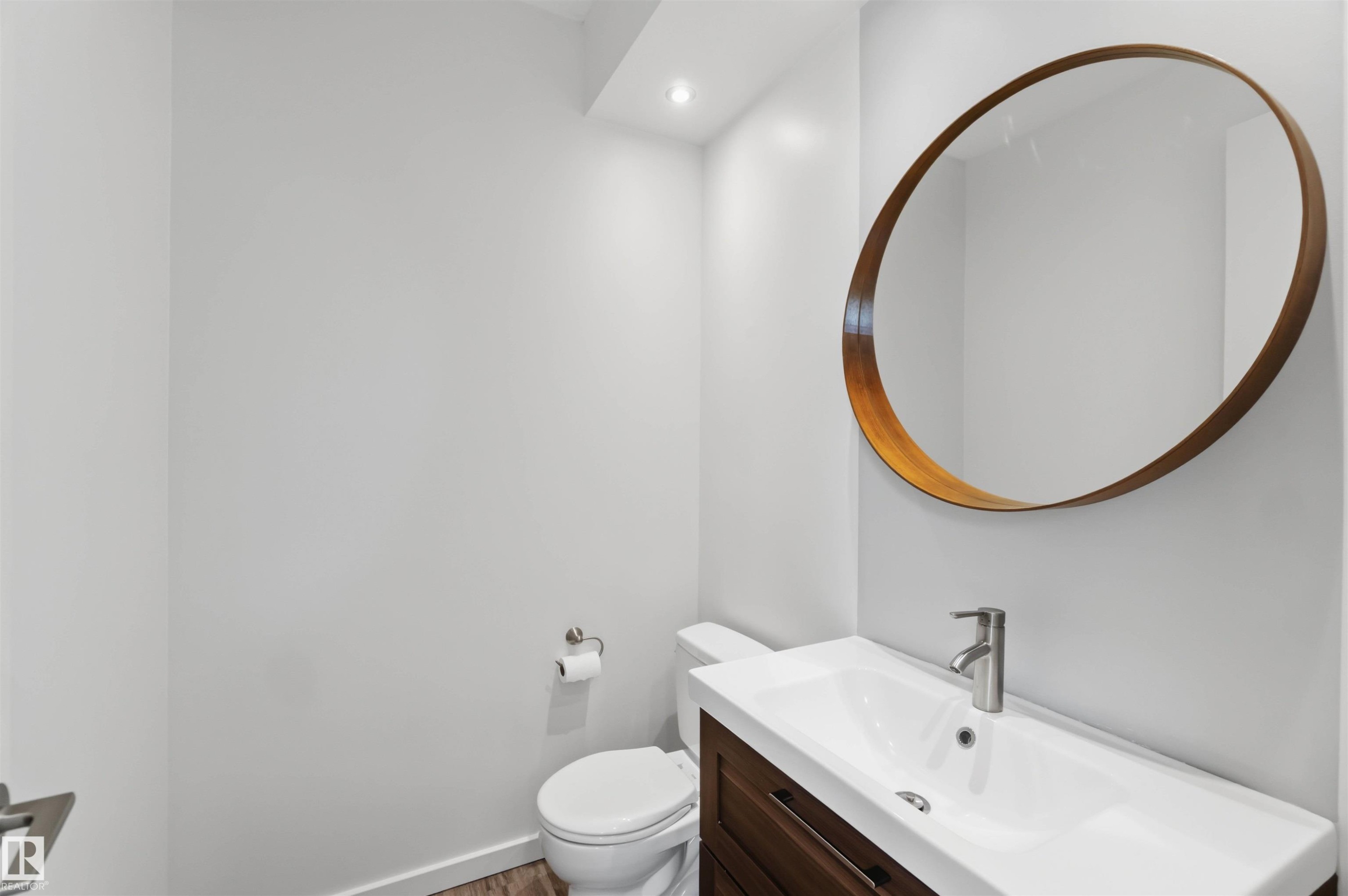 Modern powder room featuring a wall-mounted round mirror with a wood-tone frame, a rectangular vanity with a white basin sink, a brushed nickel faucet, and wood-finish flooring - #1 14315 Stony Plain Nw, Edmonton, AB - Indoor Photo Showing Bathroom