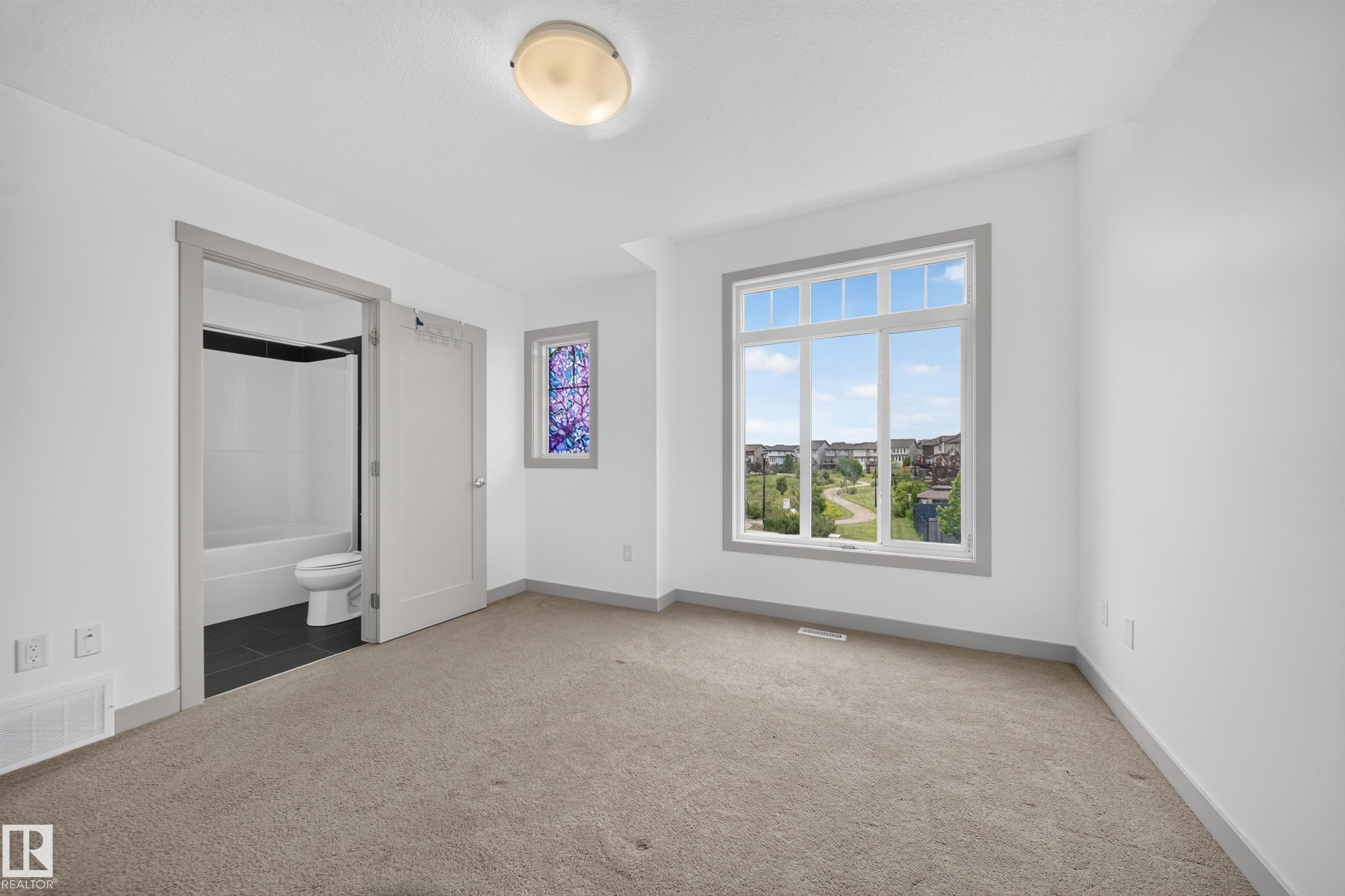 Carpeted room featuring a large window with white framing, a stained-glass accent window, and an en-suite bathroom with dark floor tiling and a white tub/shower combination - 227 Cy Becker Blvd, Edmonton, AB