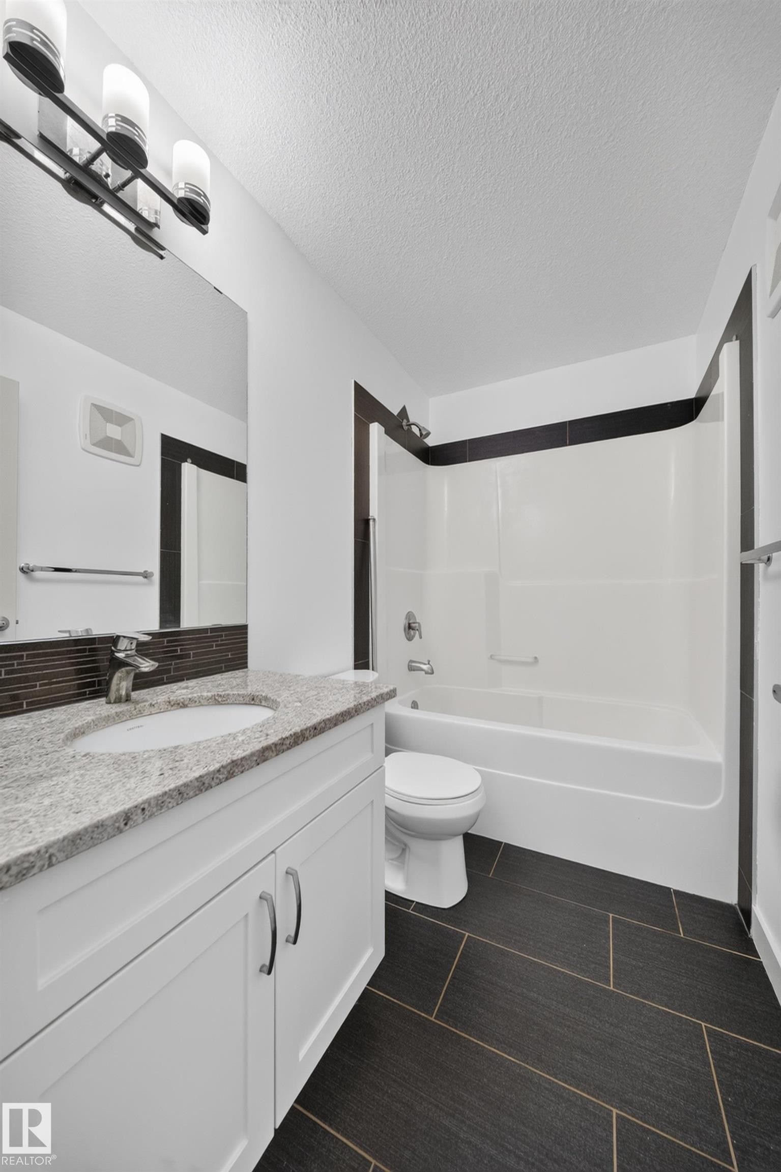 Bathroom featuring white cabinetry, a single basin sink, a speckled countertop, a shower-tub combination, and dark rectangular floor tiles - 227 Cy Becker Blvd, Edmonton, AB
