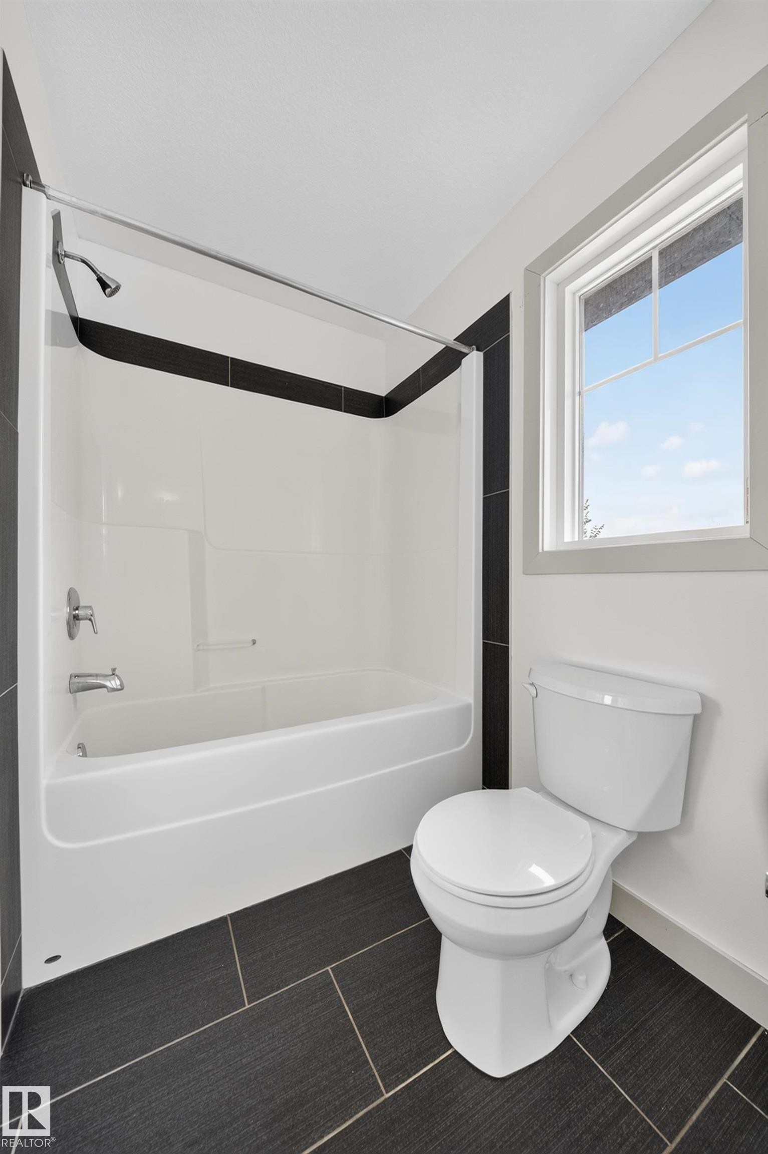 Bathroom featuring a white bathtub and shower combination with chrome fixtures, a two-piece toilet, dark large-format floor tiles, and a white framed window - 227 Cy Becker Blvd, Edmonton, AB