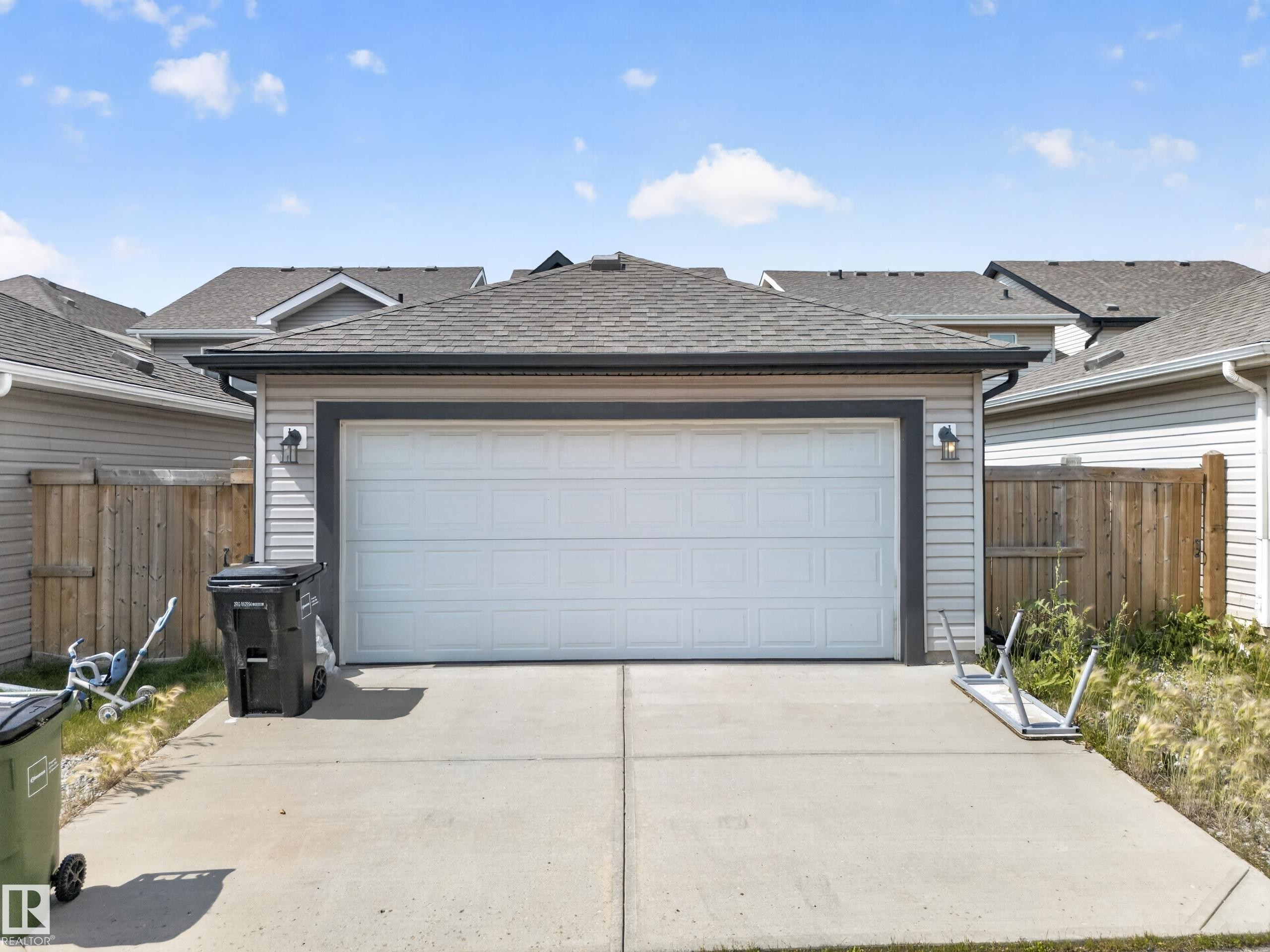 Attached garage with a white overhead door, concrete driveway, light-toned siding, dark trim, and twin exterior light fixtures - 227 Cy Becker Blvd, Edmonton, AB