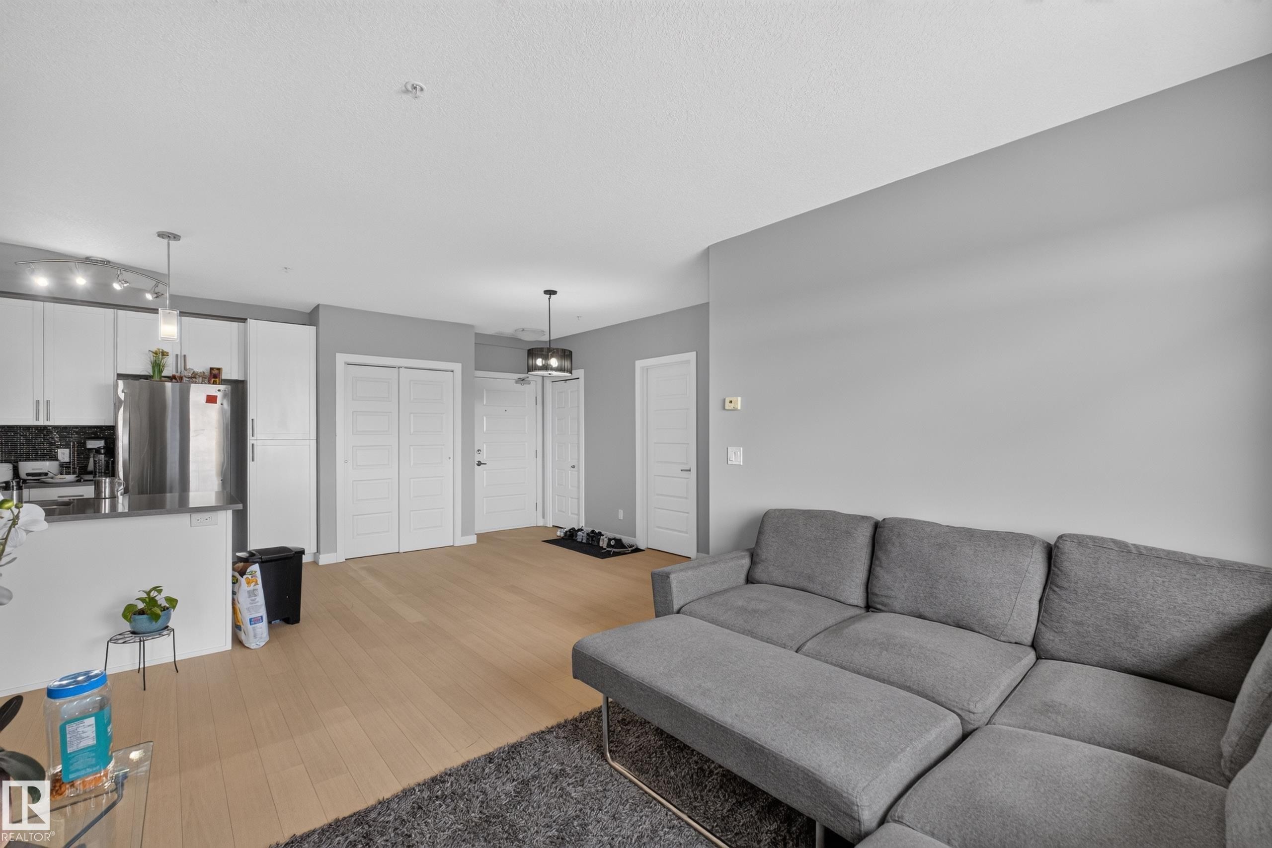 Open-concept living area featuring light wood-finish flooring, a modern kitchen with stainless steel appliances, white cabinetry, and pendant lighting - 706 5151 Windermere Boulevard, Edmonton, AB