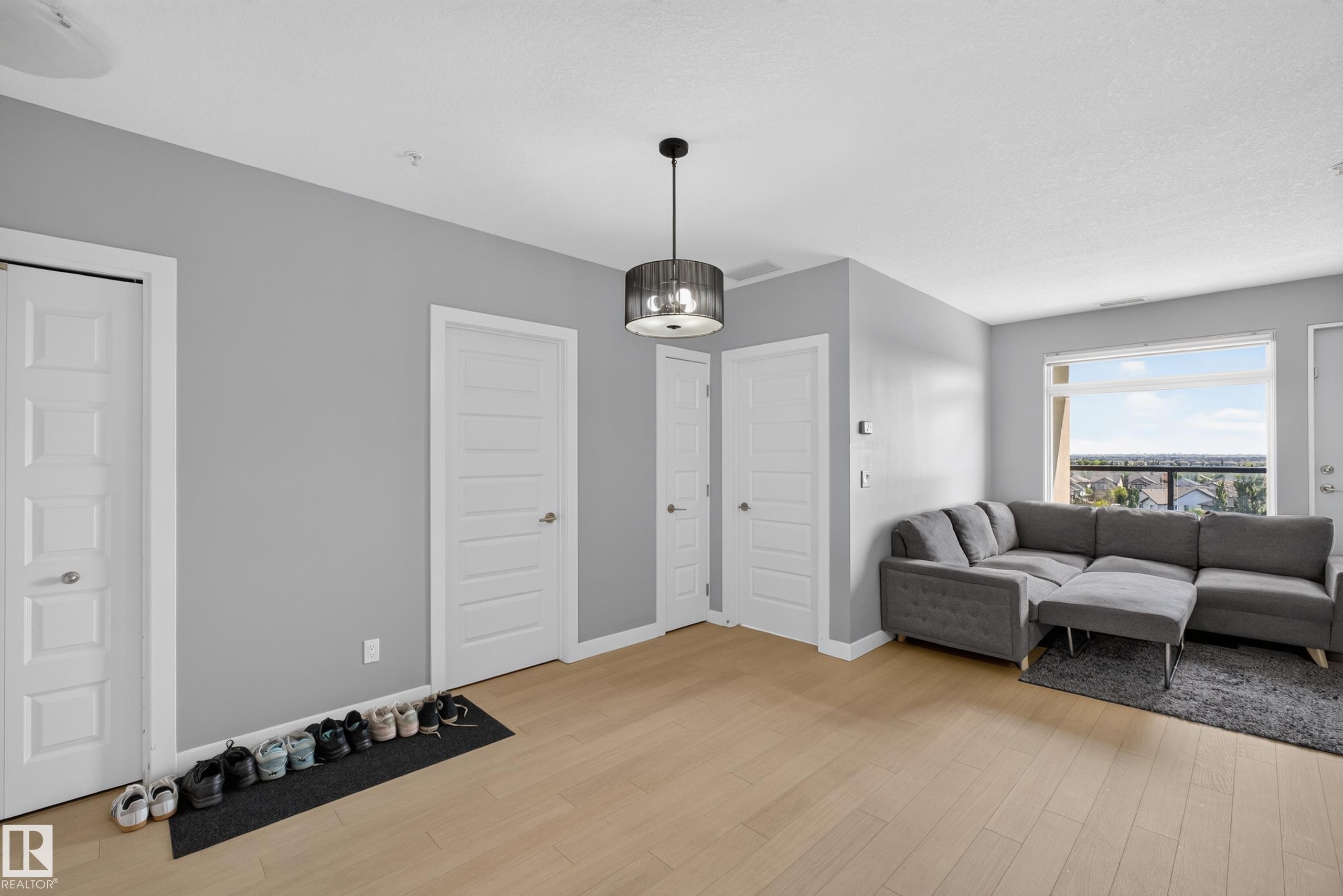 Open-concept living area featuring light wood-finish flooring, a contemporary ceiling light fixture, a large window offering expansive outdoor scenery, and neutral gray wall paint - 706 5151 Windermere Boulevard, Edmonton, AB