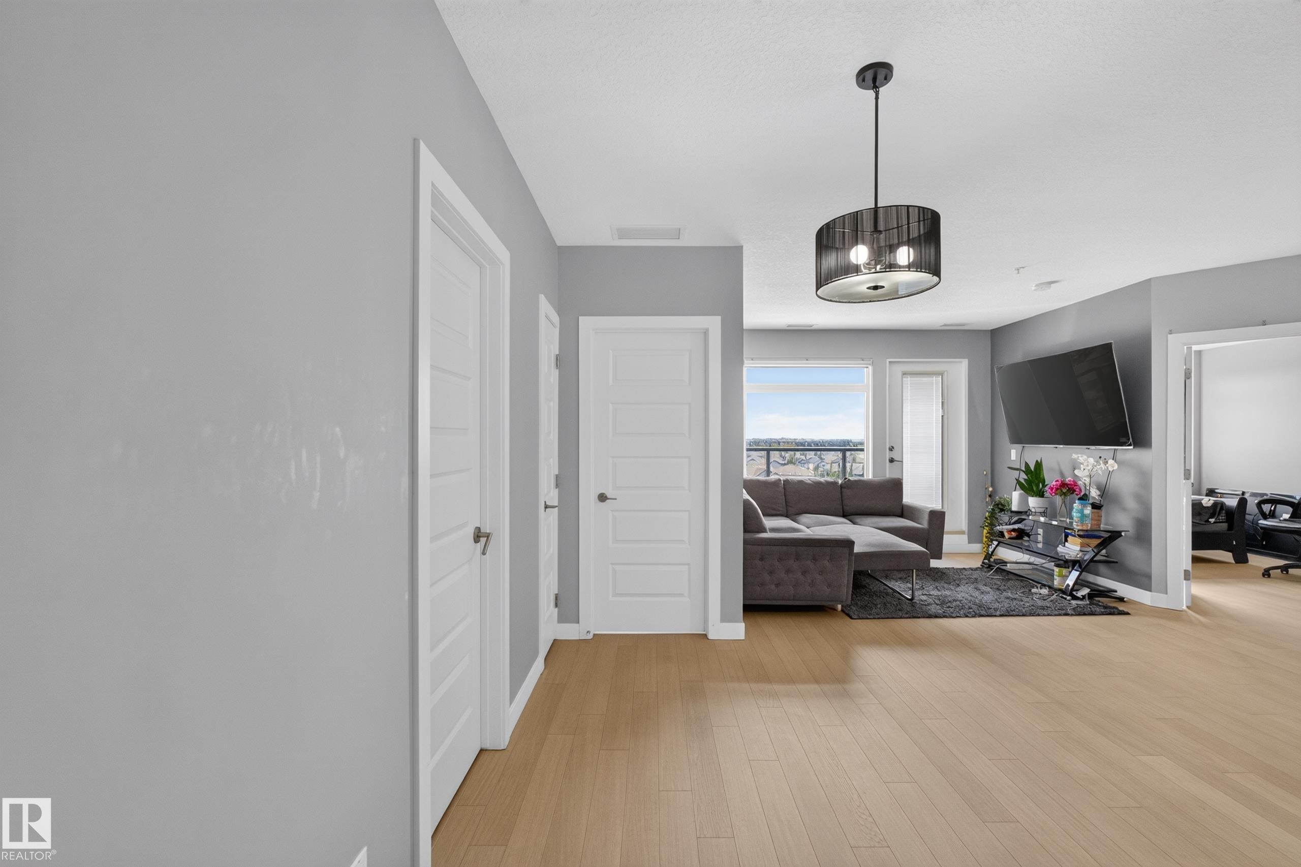 Open-concept living area featuring wood-finish flooring, a modern drum pendant light fixture, and light gray wall tones - 706 5151 Windermere Boulevard, Edmonton, AB