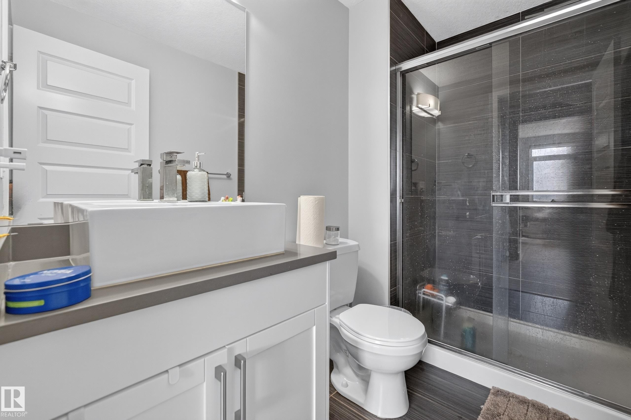 Bathroom featuring a glass-enclosed shower with dark tile surround, a vessel sink on a vanity with white cabinetry, and wood-finish flooring - 706 5151 Windermere Boulevard, Edmonton, AB
