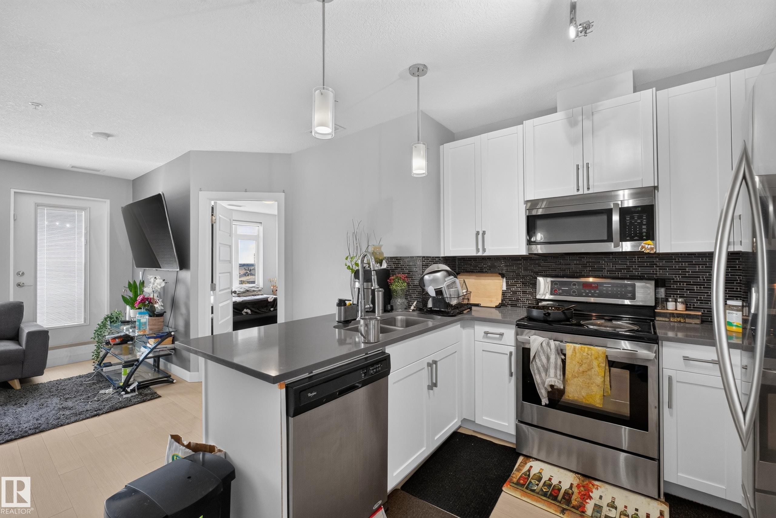 Modern kitchen featuring white cabinetry, stainless steel appliances, a dark tile backsplash, and light wood-finish flooring - 706 5151 Windermere Boulevard, Edmonton, AB