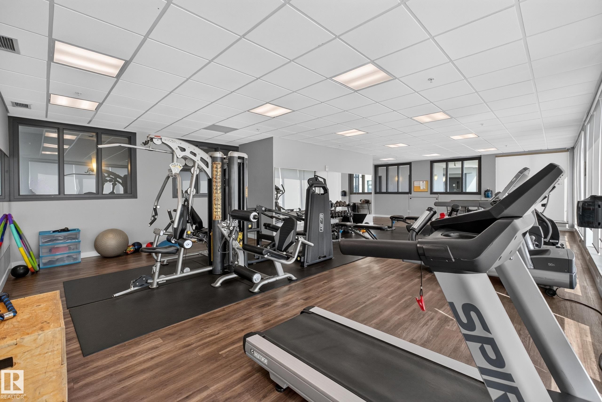 Spacious fitness room featuring wood-finish flooring, drop ceiling with recessed lighting, large windows, and mirrored walls - 706 5151 Windermere Boulevard, Edmonton, AB