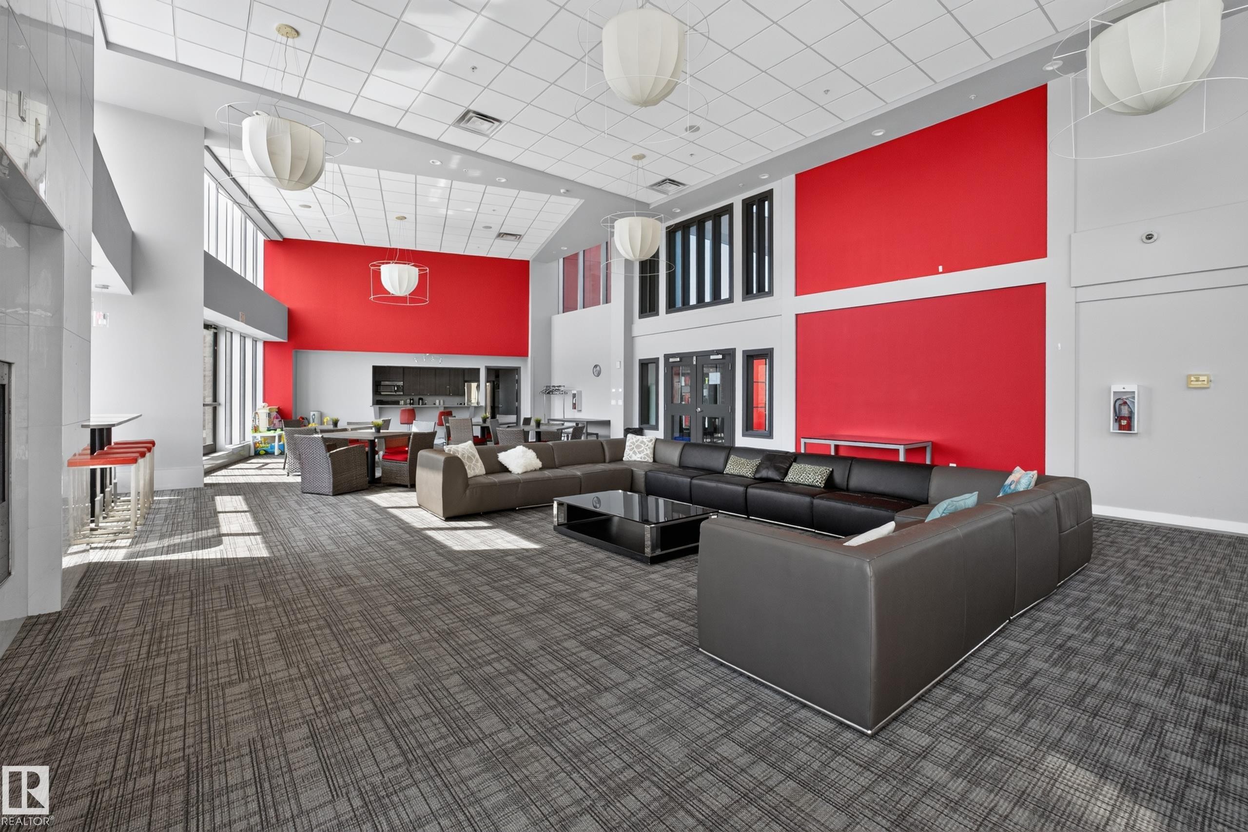 Spacious lounge area featuring high ceilings, contemporary pendant lighting, patterned carpet flooring, a large window wall, and vibrant accent walls - 706 5151 Windermere Boulevard, Edmonton, AB