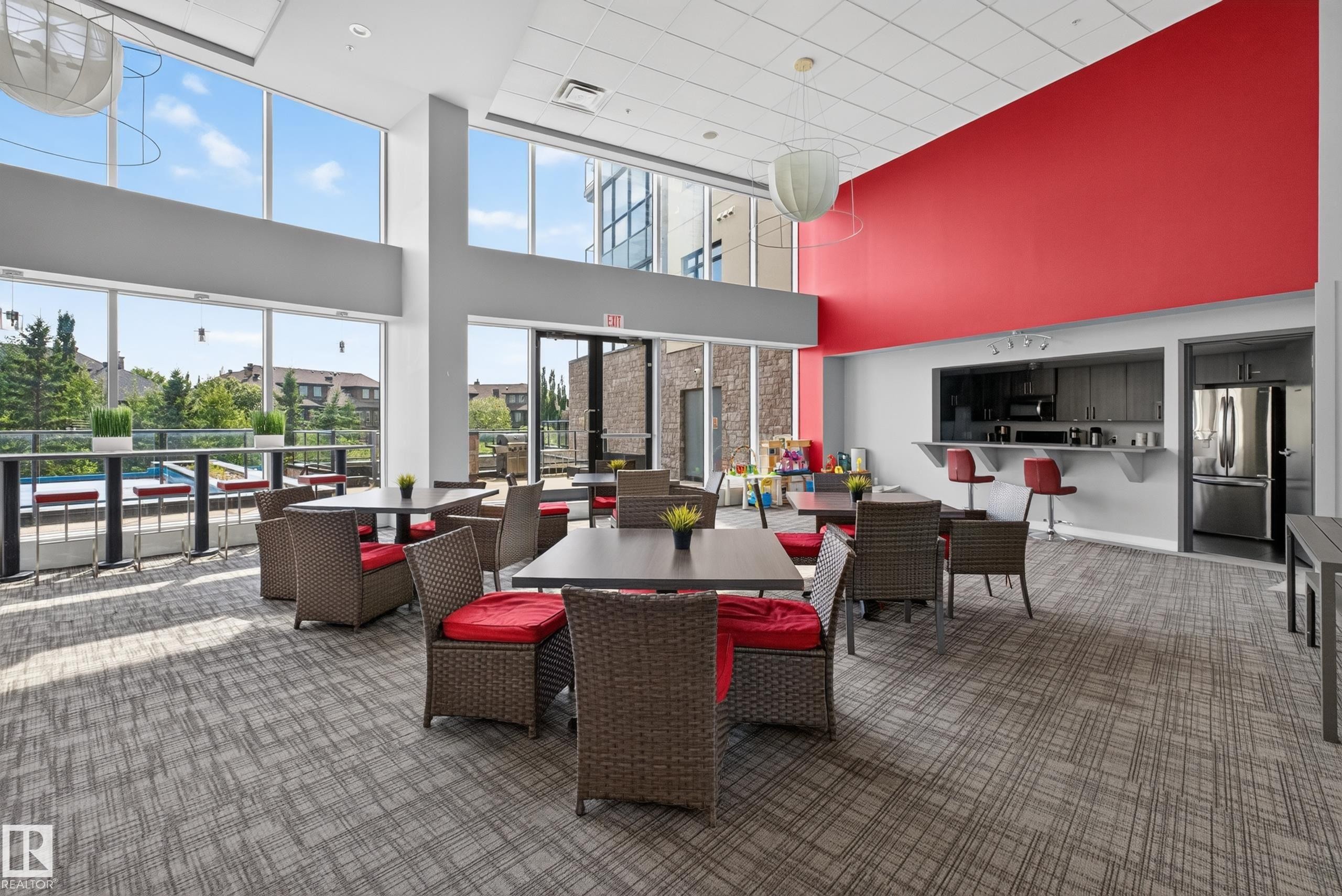 Spacious common area featuring large windows, a full-height red accent wall, and a built-in wet bar with gray cabinetry and stainless steel appliances - 706 5151 Windermere Boulevard, Edmonton, AB