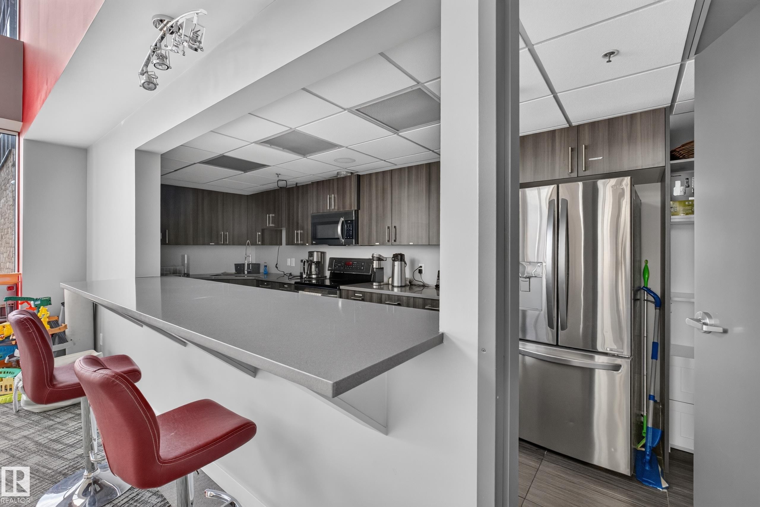 Contemporary kitchen with a pass-through bar, featuring dark wood-finish cabinetry, stainless steel appliances, and a gray countertop - 706 5151 Windermere Boulevard, Edmonton, AB