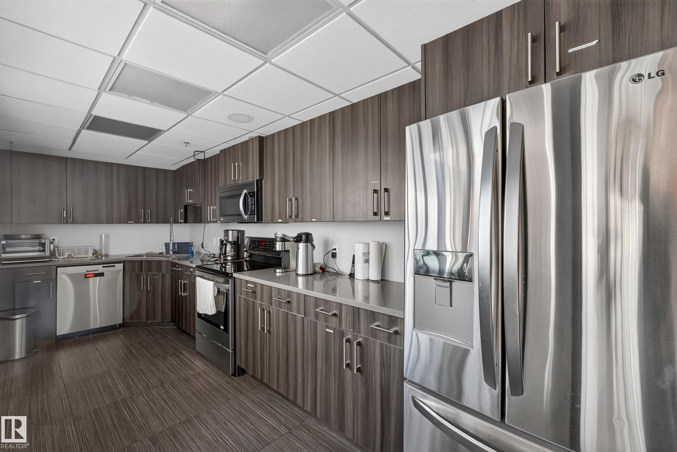 Modern kitchen featuring wood-finish cabinetry, stainless steel appliances, and a drop ceiling with integrated lighting - 706 5151 Windermere Boulevard, Edmonton, AB