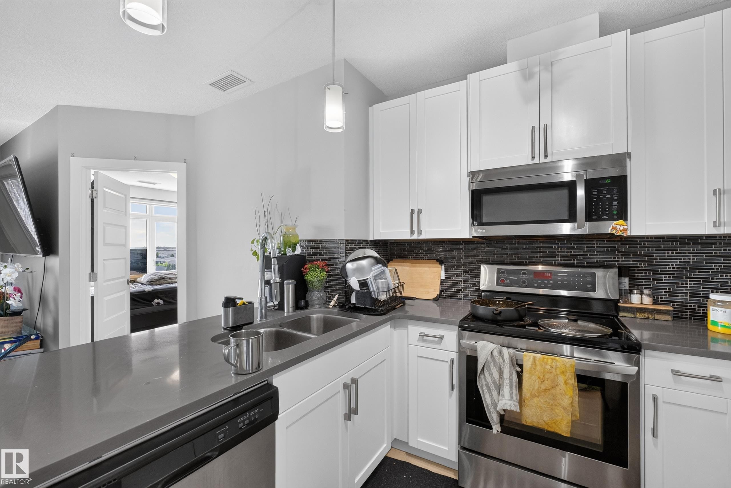 Modern kitchen featuring white shaker cabinetry, stainless steel appliances, a dark tile backsplash, and light gray countertops - 706 5151 Windermere Boulevard, Edmonton, AB