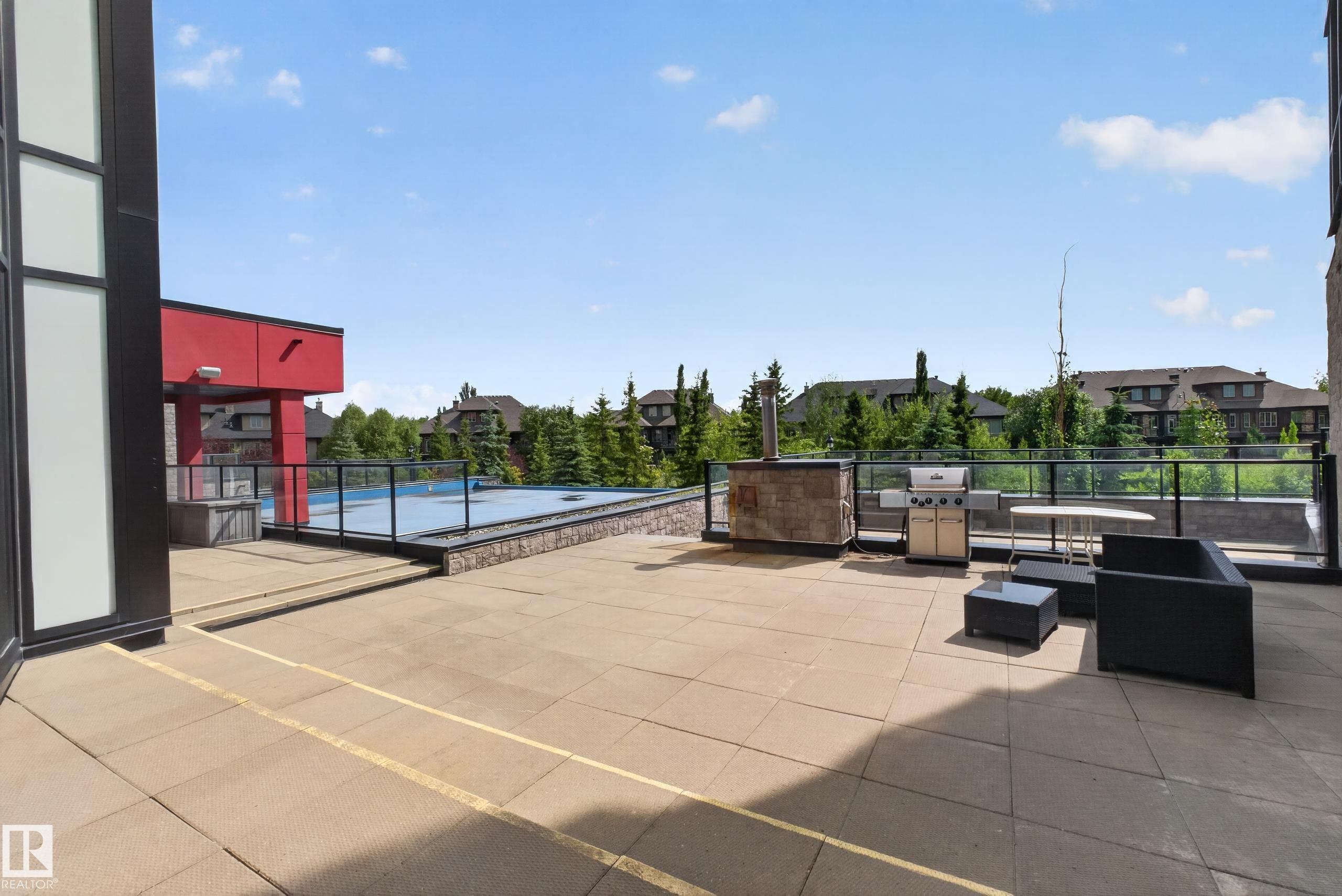 Outdoor paved patio area featuring a stone-clad built-in barbecue station, glass railing, and a visible swimming pool - 706 5151 Windermere Boulevard, Edmonton, AB