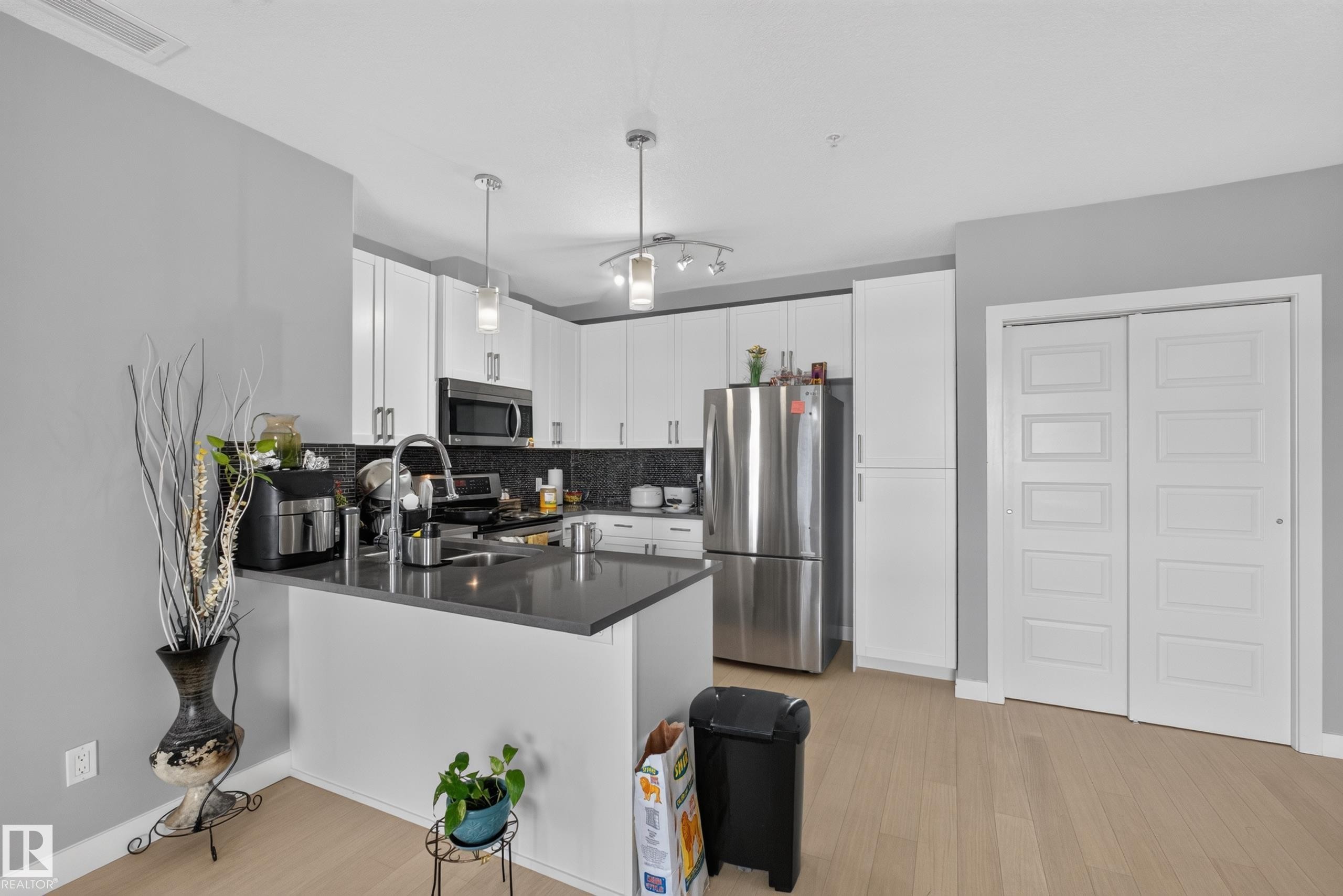 Modern kitchen featuring white cabinetry, stainless steel appliances, a dark countertop, a dark patterned backsplash, and wood-finish flooring - 706 5151 Windermere Boulevard, Edmonton, AB